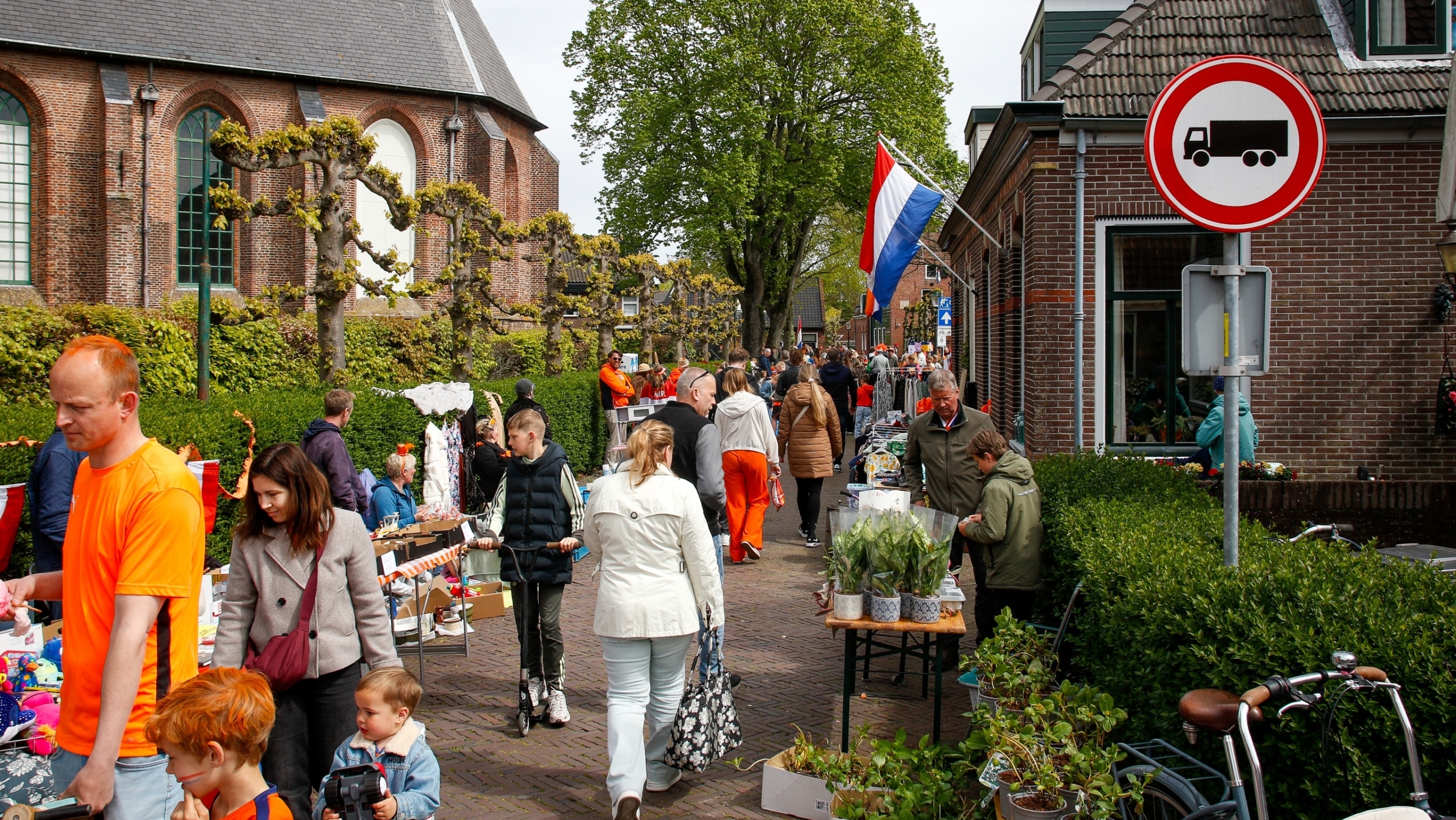 Mensen lopen langs een drukke markt met kraampjes en een Nederlandse vlag, nabij een historische kerk en gebouwen.