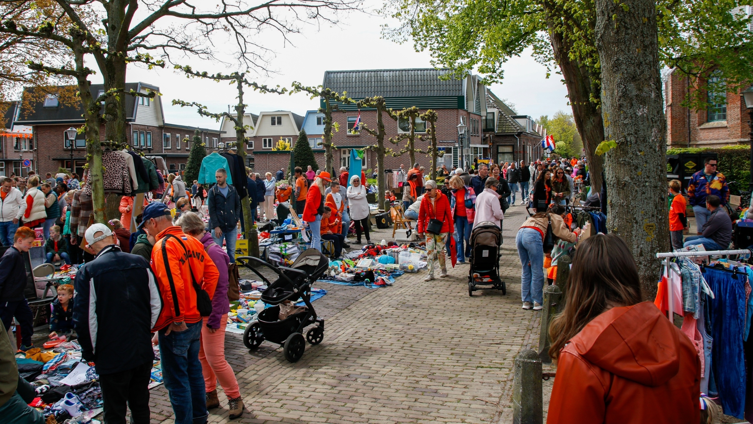 Mensen wandelen en bekijken kraampjes bij een vrijmarkt op een zonnige dag, omgeven door bomen en huizen.