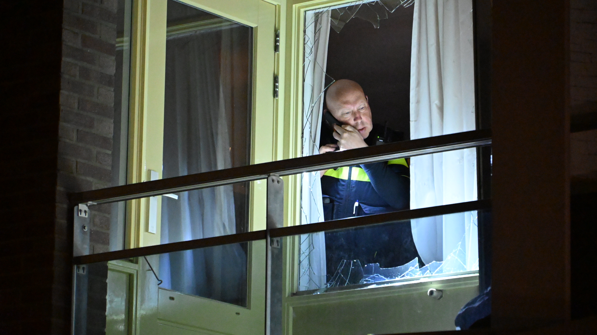 Politieagent aan de telefoon bij een kapot raam in een gebouw, gefotografeerd in het donker.