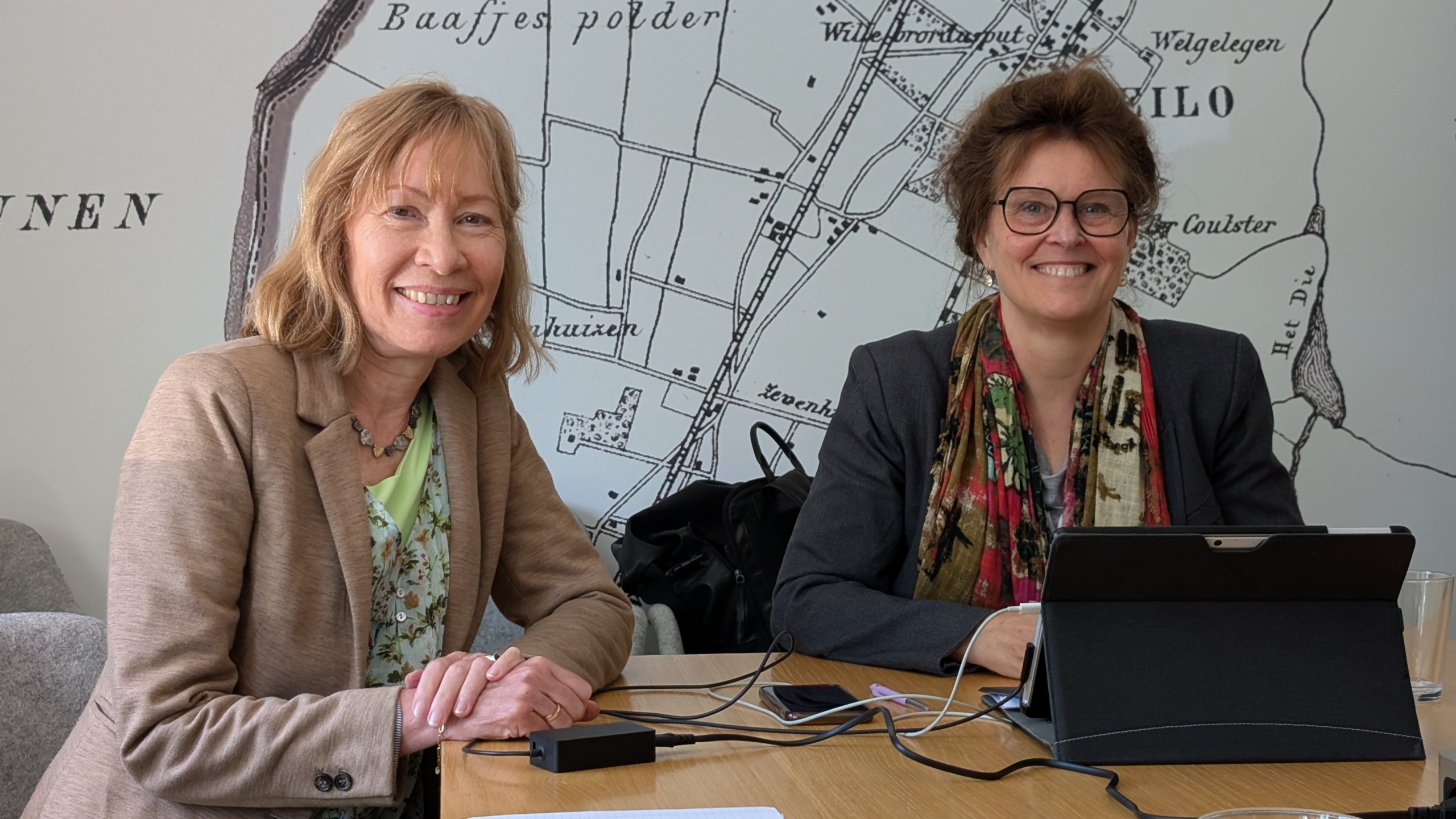 Twee lachende vrouwen zitten aan een tafel met een laptop, tegen een achtergrond van een oude kaart.