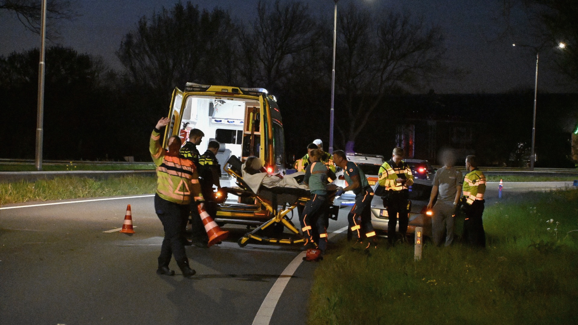 Hulpdiensten bij een ambulance op een weg in de nacht, omringd door bomen en straatverlichting.
