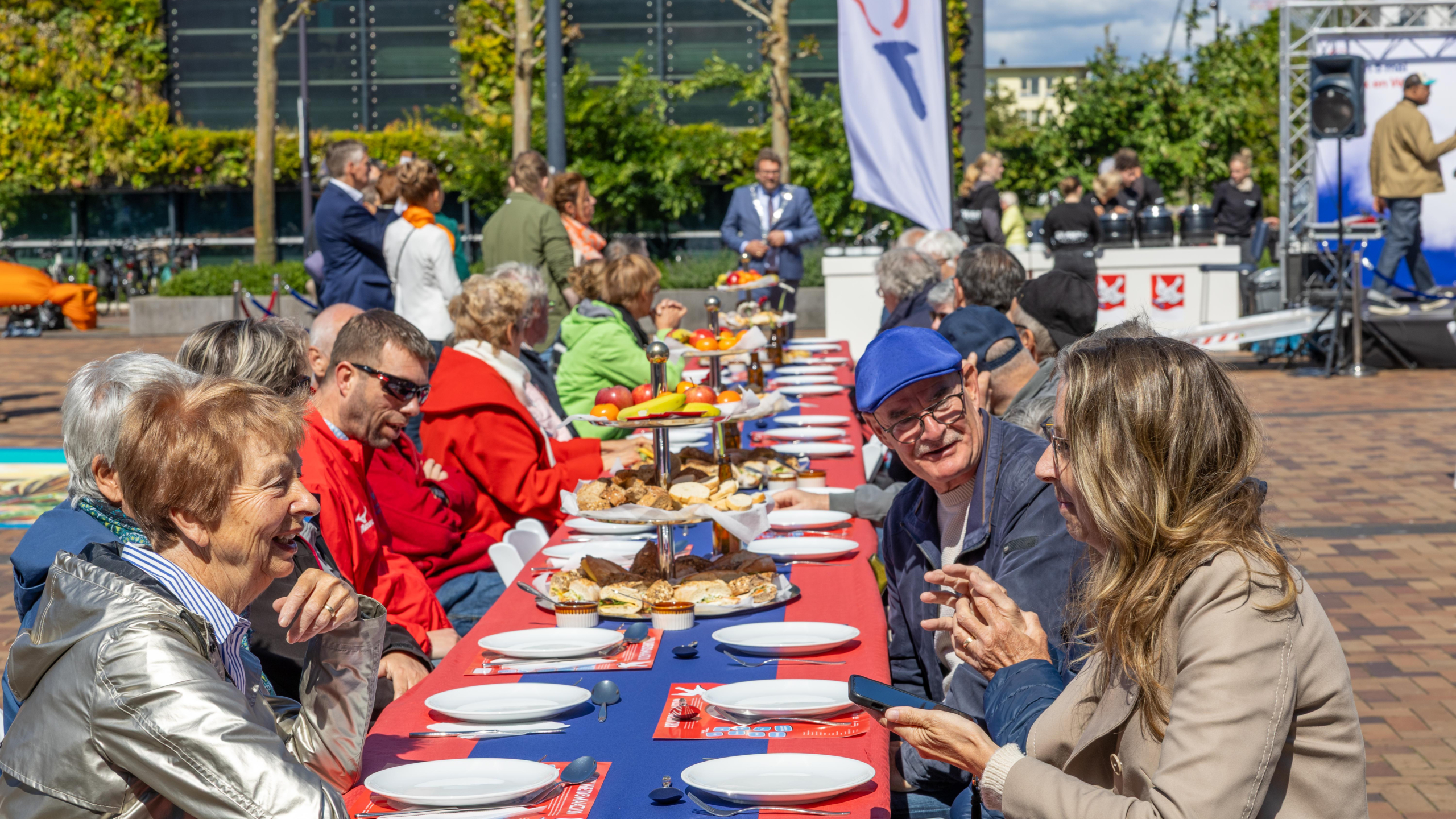 Mensen zitten aan lange gedekte tafels in de buitenlucht tijdens een evenement, met kosteloos eten en drinken.