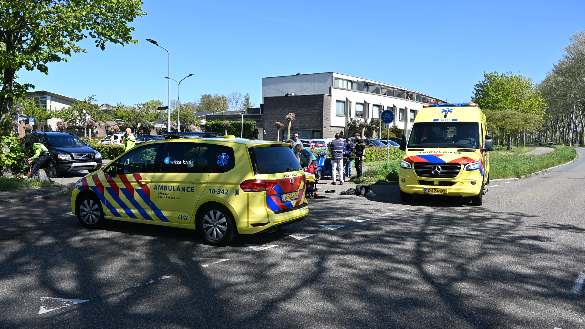 Twee ambulances staan op straat bij een verkeersongeval, met hulpverleners die zich bezighouden met een motorfiets en andere voertuigen.