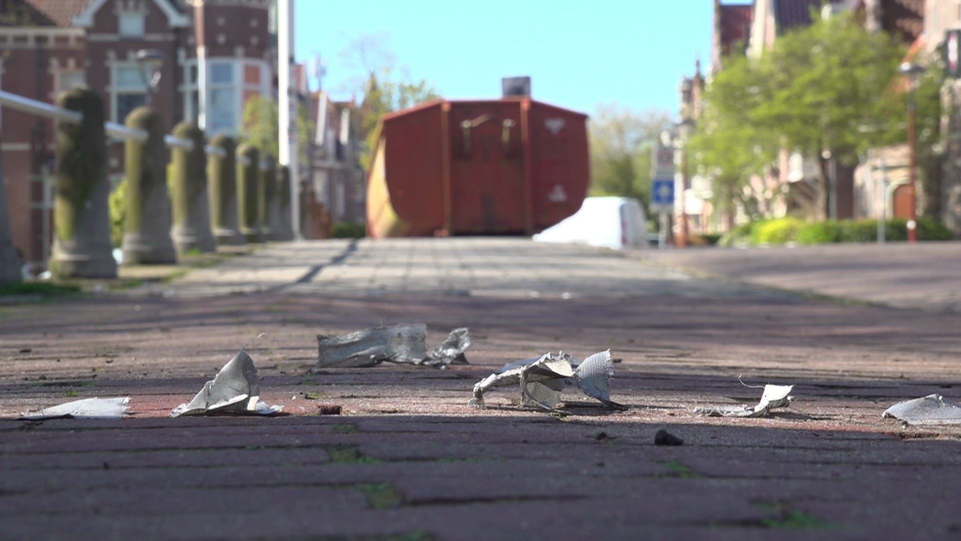 Stukken aluminiumfolie liggen verspreid over de stoep, met op de achtergrond een grote rode container op een rustige straat.