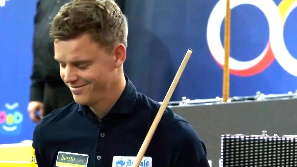 Man in een donkere outfit met een glimlach, houdend een biljartkeu, tegen een achtergrond met een logo.