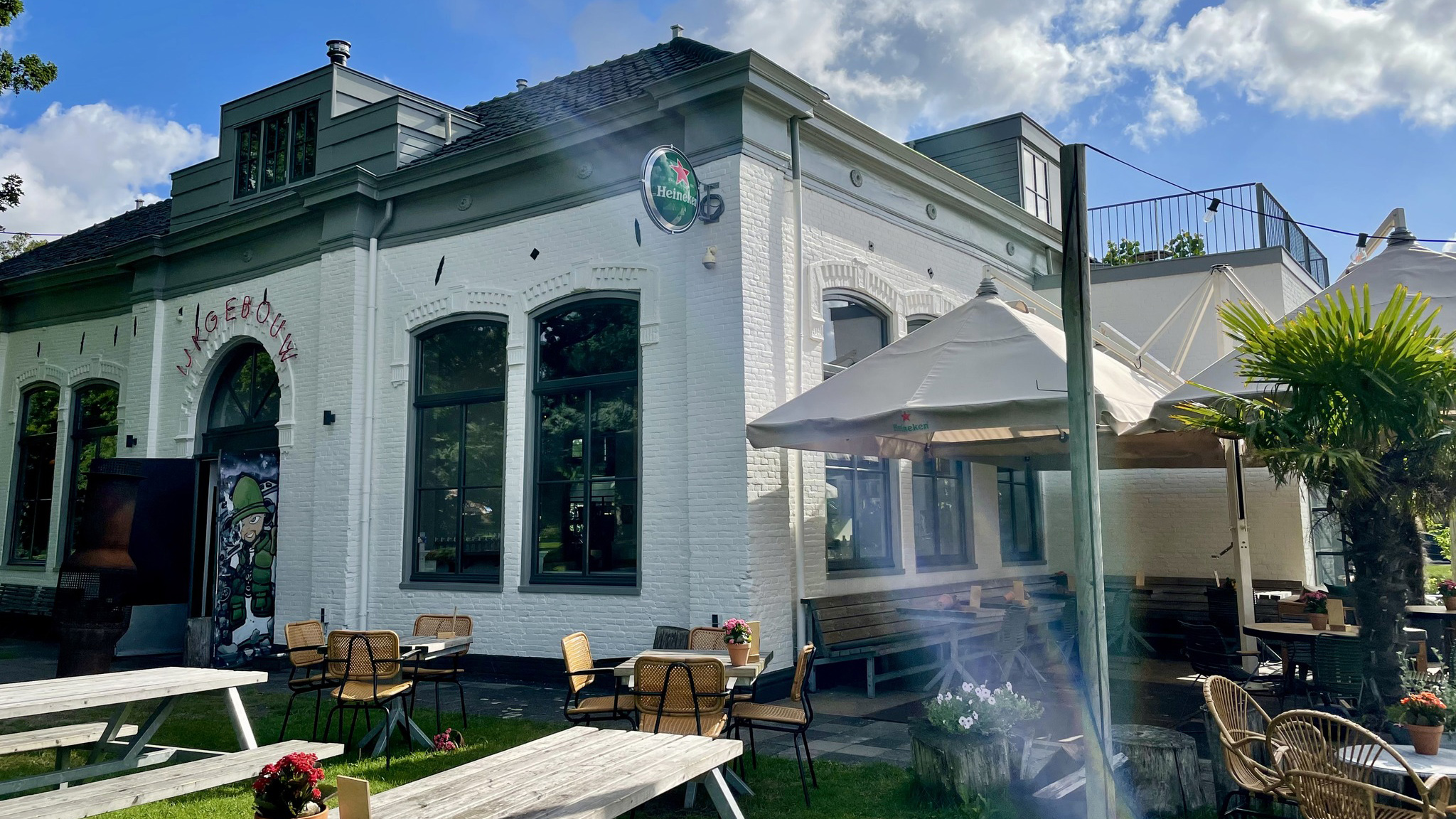 Wit gebouw met terras, parasols, en tuinmeubilair, omringd door bloemen en groen onder een blauwe hemel.