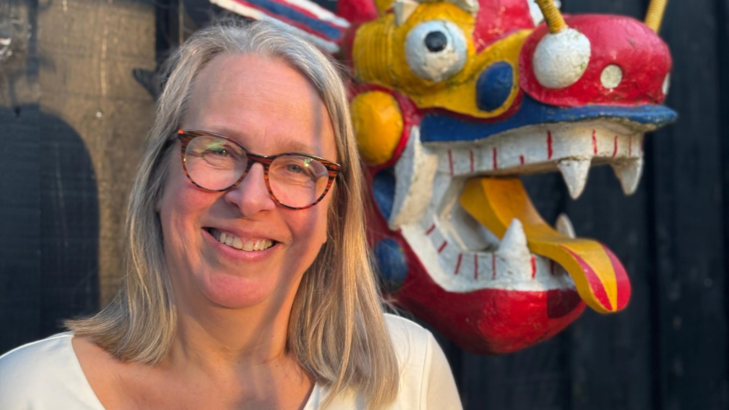 Vrouw met grijs haar en bril staat glimlachend naast een kleurrijk beeld van een draak met grote ogen en tanden.