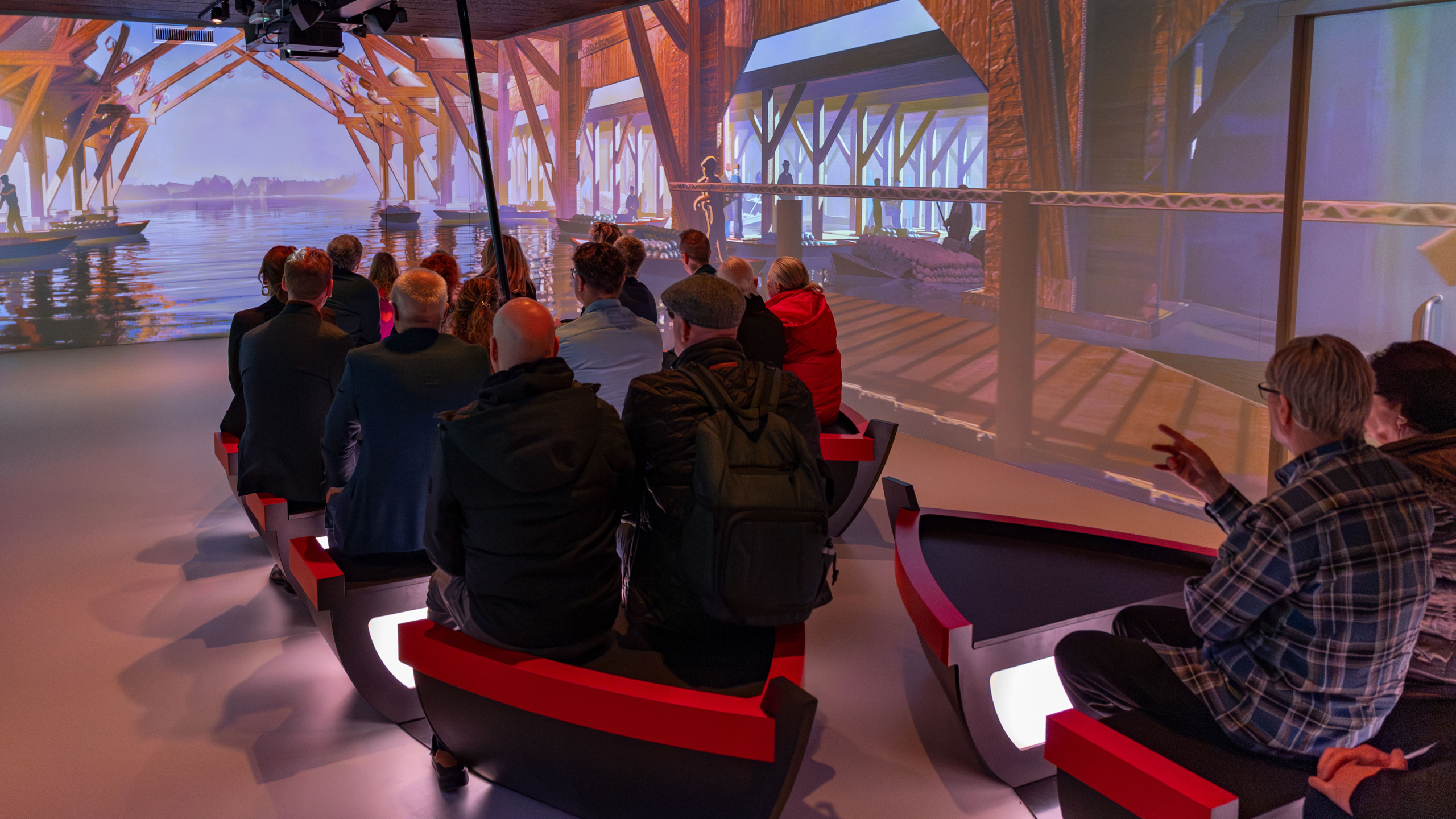 Mensen zitten op moderne stoelen en kijken naar een grote projectie van een houten structuur en waterlandschap.