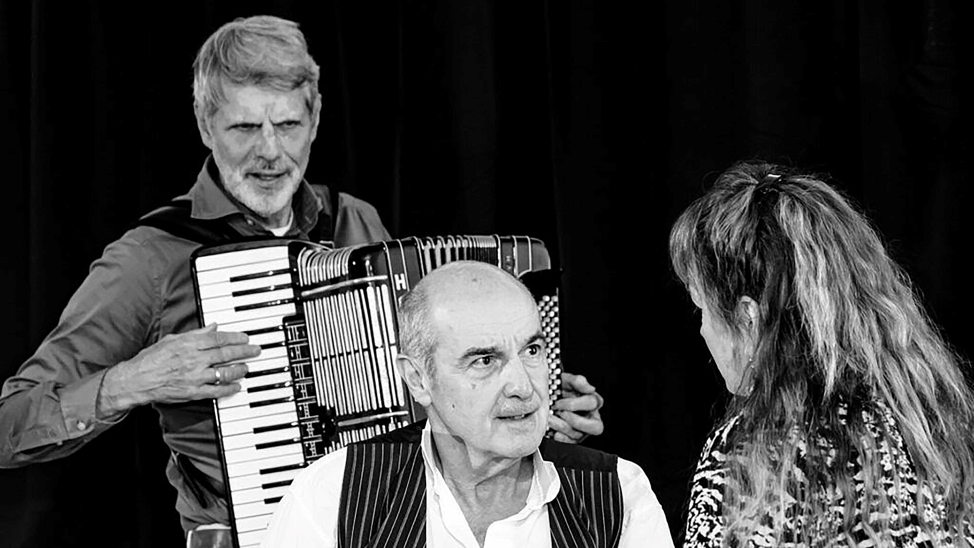Twee mannen en een vrouw in gesprek, met een man die een accordeon bespeelt, gefotografeerd in zwart-wit.