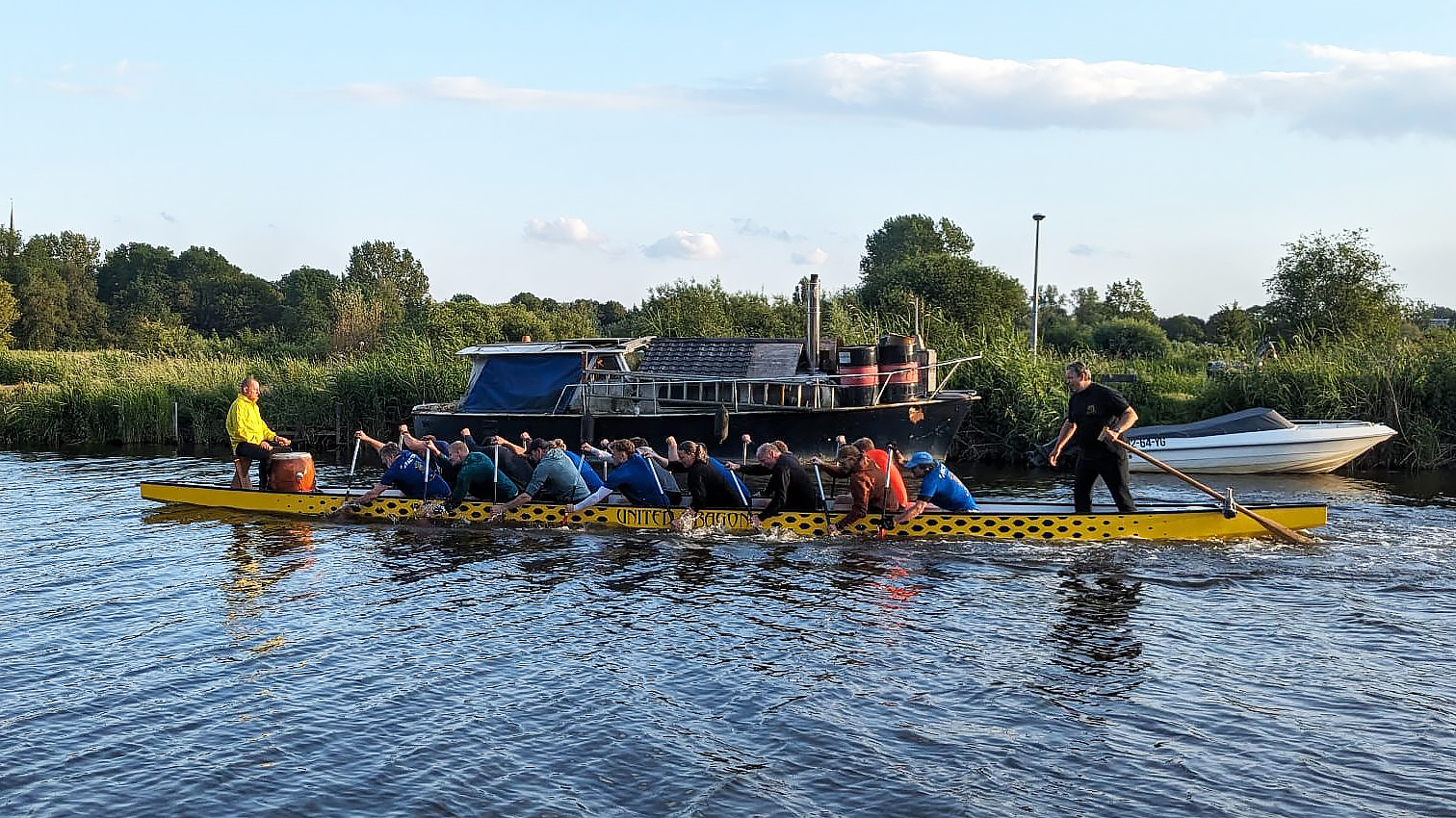 Mensen roeien in een gele drakenboot op een rivier, met groene oevers en boten op de achtergrond.