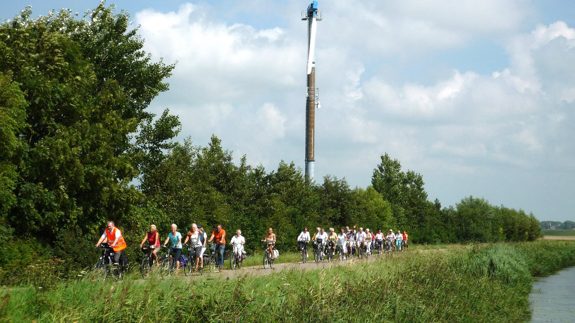 Grote groep fietsers rijdt langs een groen pad met bomen en een hoge toren op de achtergrond.