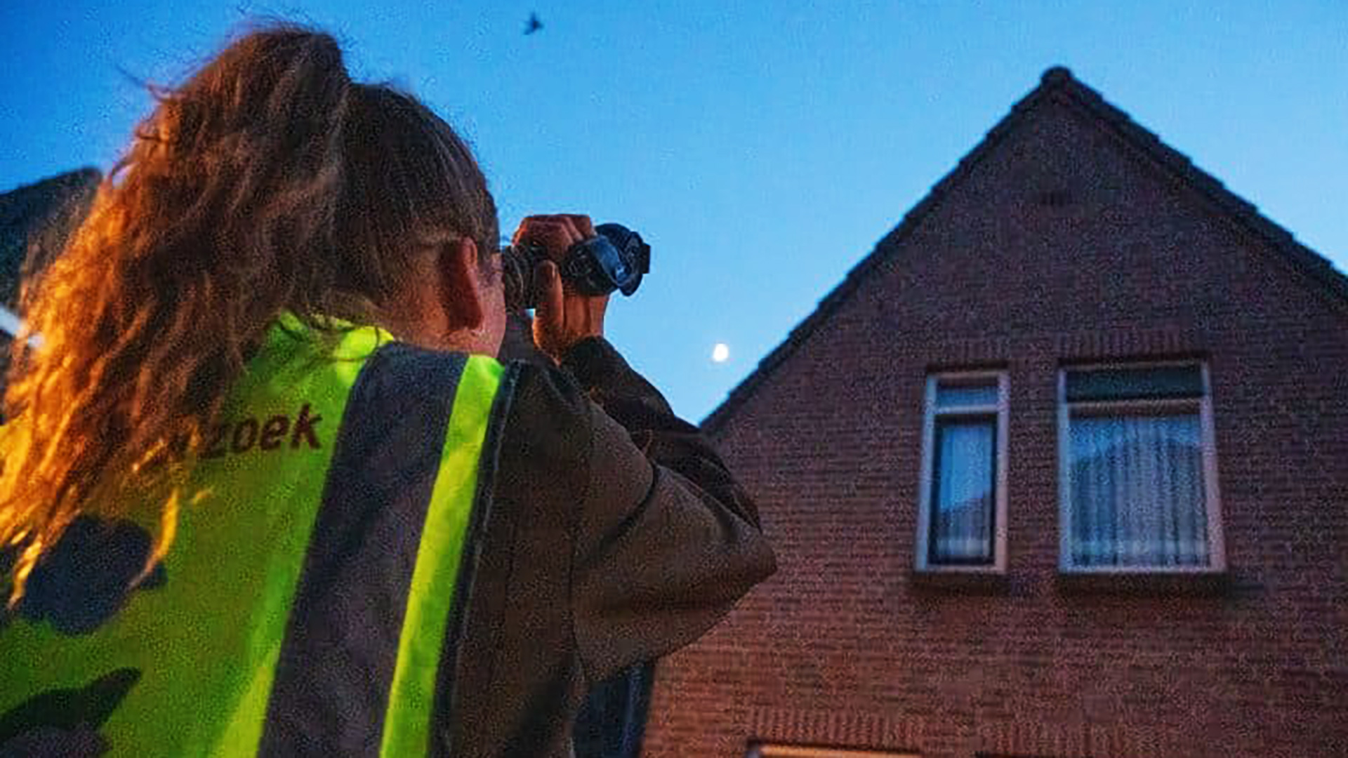 Een persoon in een reflecterend vest kijkt 's avonds met een verrekijker naar een bakstenen huis en de lucht.