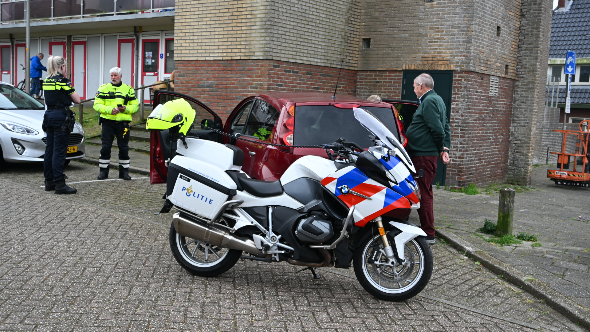 Agenten praten met een man naast een rode auto; een politiefiets met blauwe en rode strepen staat op de voorgrond.
