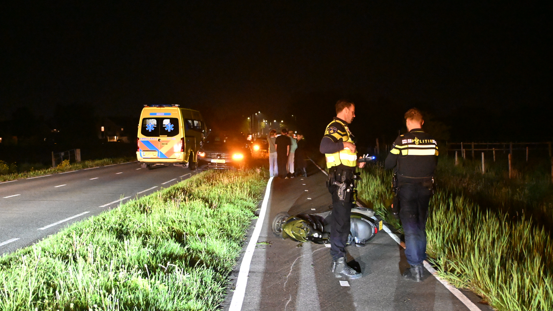 Twee politieagenten en een omgevallen scooter langs de weg bij nacht, met een ambulance op de achtergrond.