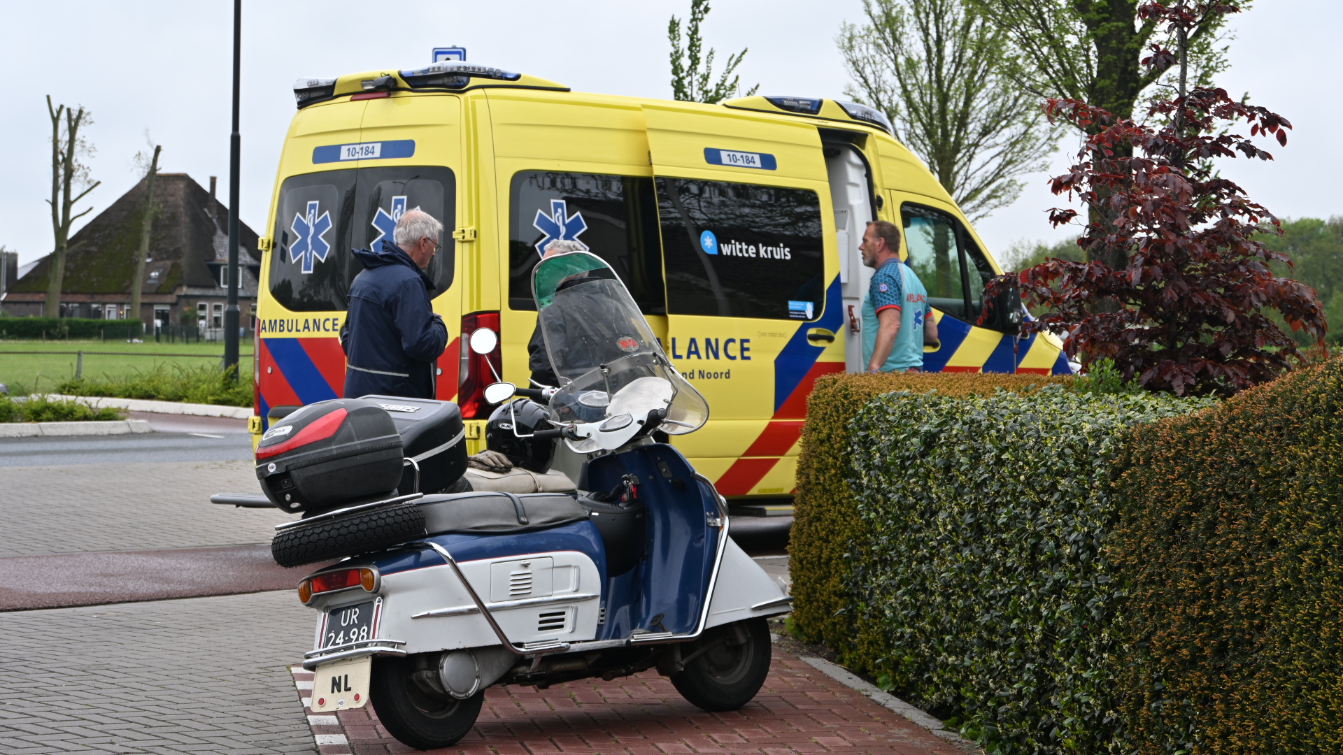 Gele ambulance met mensen ernaast en een geparkeerde scooter op een straat, met groene heg op de voorgrond.