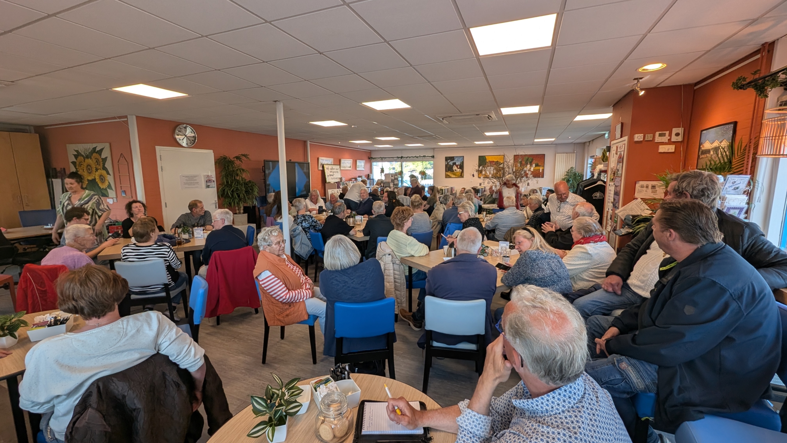 Volle gemeenschapsruimte met mensen die aan tafels zitten en praten, met kunstwerken aan de muren.