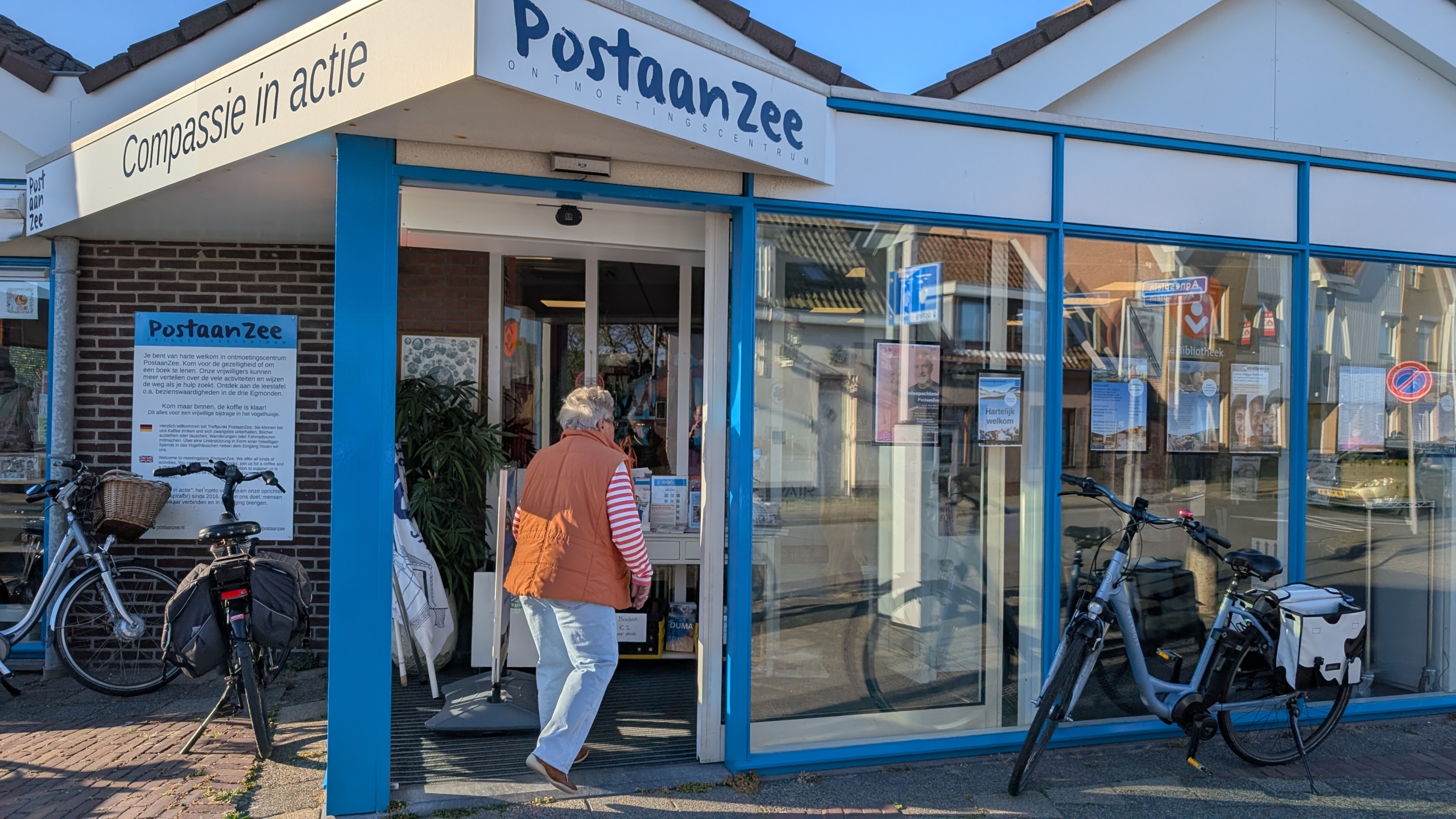 Een persoon loopt een ontmoetingscentrum genaamd "PostaanZee" binnen, met fietsen geparkeerd voor de ingang.