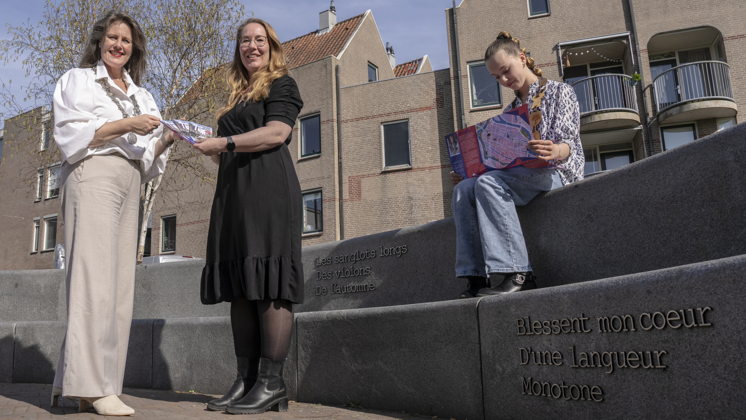 Drie mensen staan en zitten op een betonnen bank met tekst, buiten bij een gebouw, terwijl ze folders vasthouden.