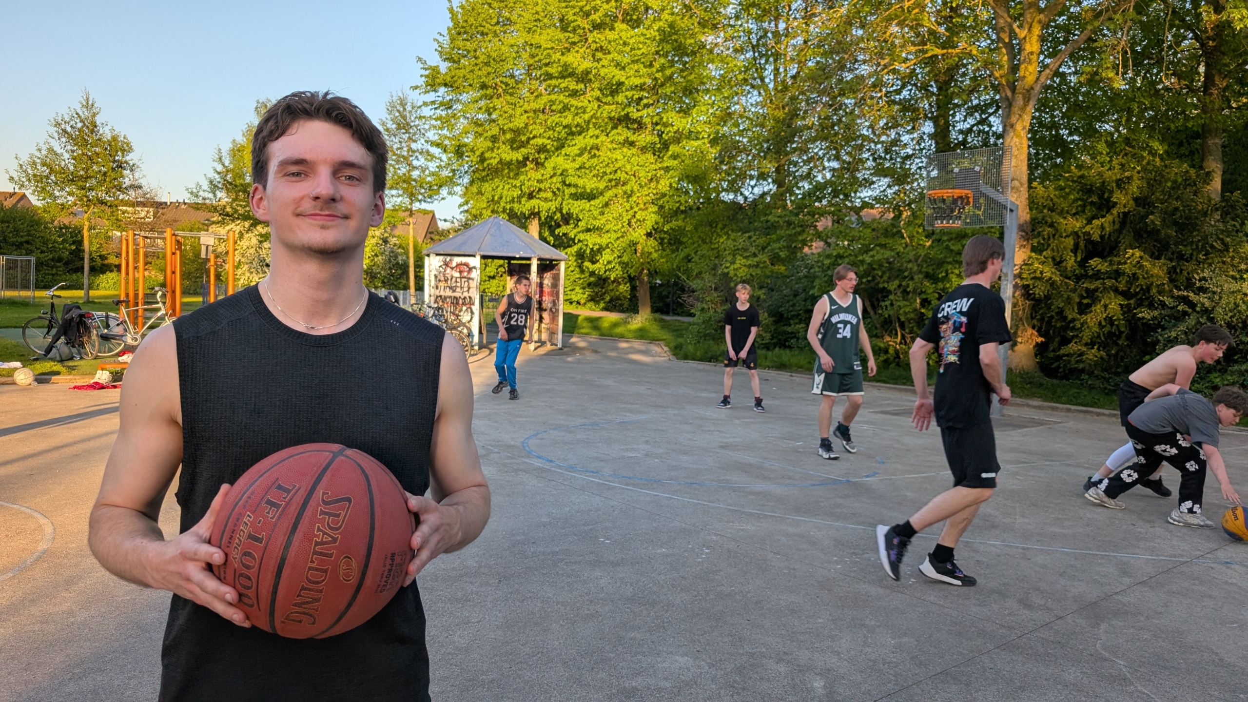 Jonge man met een basketbal op een buitenbasketbalveld, terwijl andere mensen in de achtergrond basketballen.