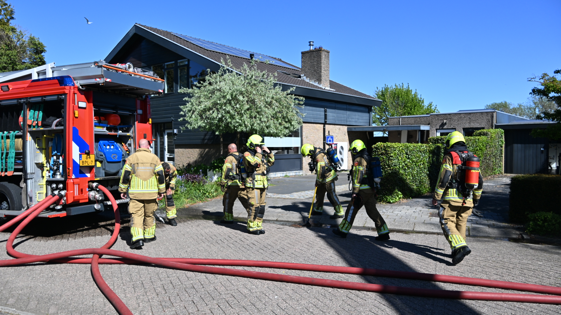 Brandweerlieden in beschermende uitrusting staan bij een brandweerwagen met uitgerolde slangen voor een modern huis.