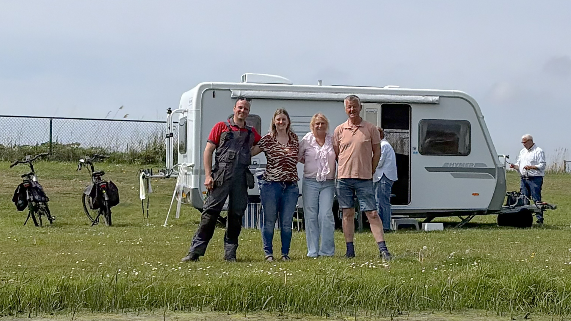 Vier mensen staan voor een caravan op een grasveld met fietsen op de achtergrond en een helderblauwe lucht.