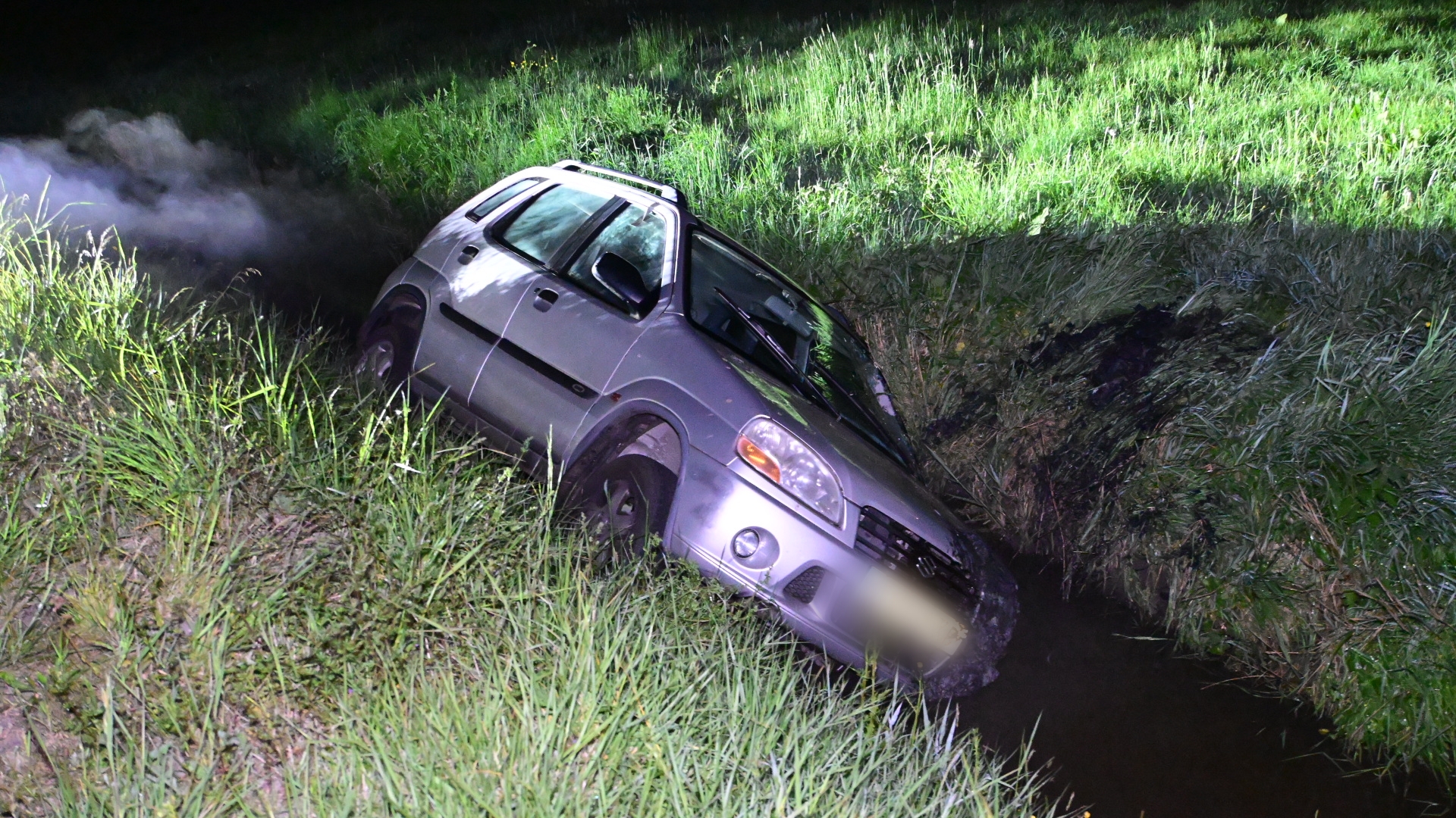 Een auto ligt gekanteld in een sloot in een grasrijke omgeving, met rook die uit de achterkant omhoog komt.