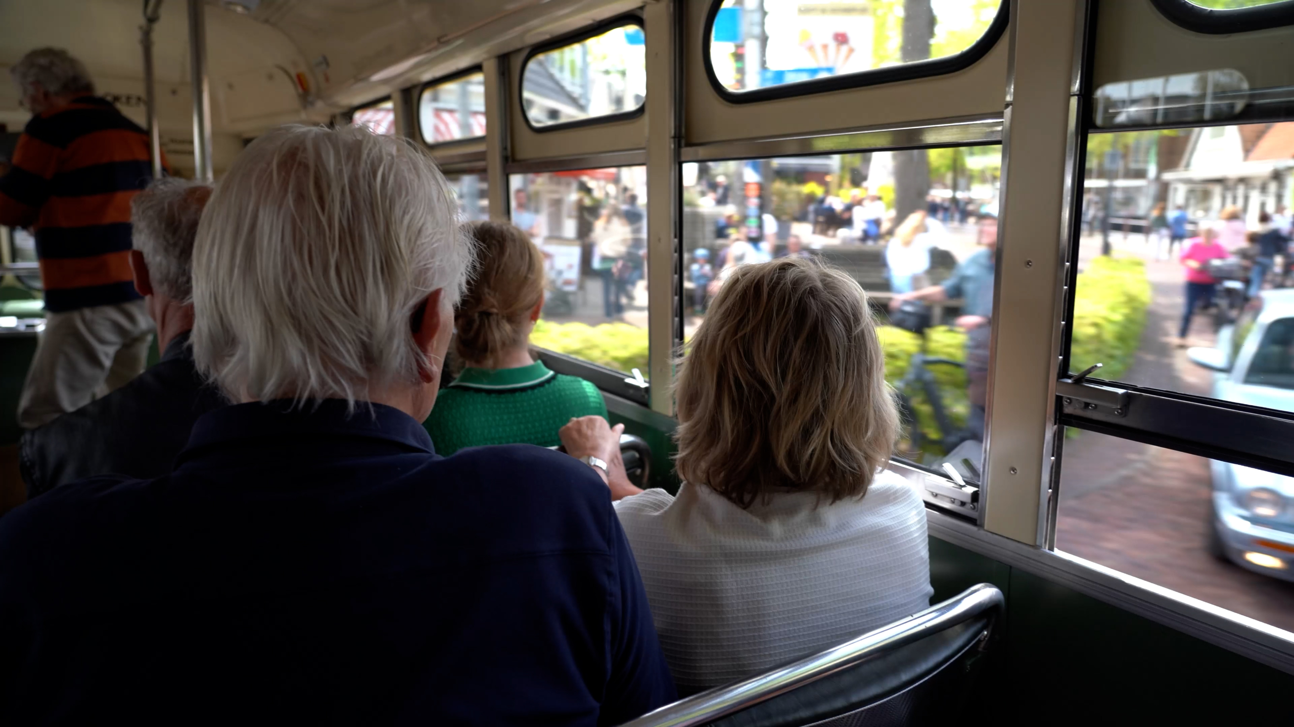 Mensen zitten in een ouderwetse bus en kijken uit het raam naar een drukke straat met voorbijgangers.