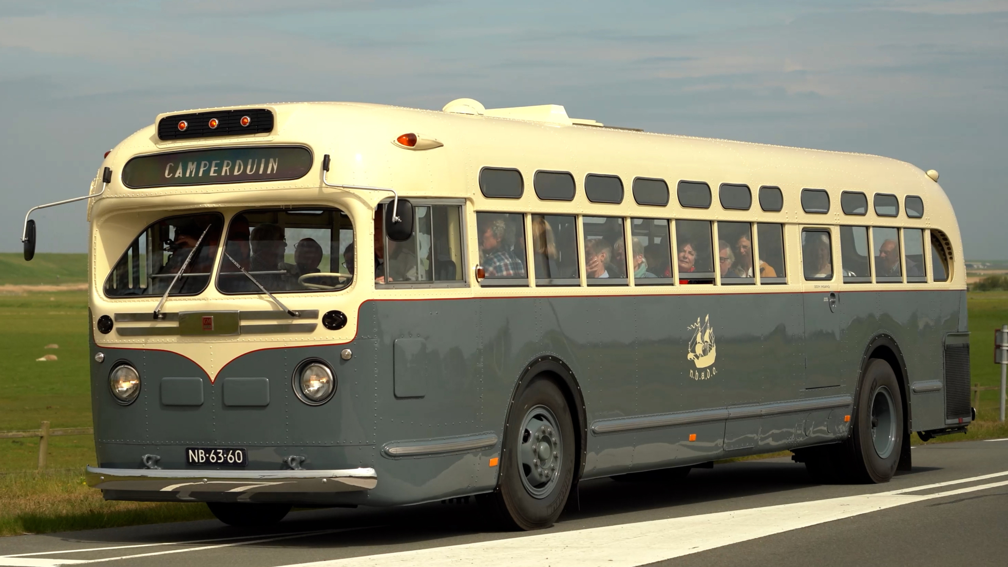 Vintage bus met opschrift "Camperduin" rijdt op een landelijke weg met groen grasland op de achtergrond.