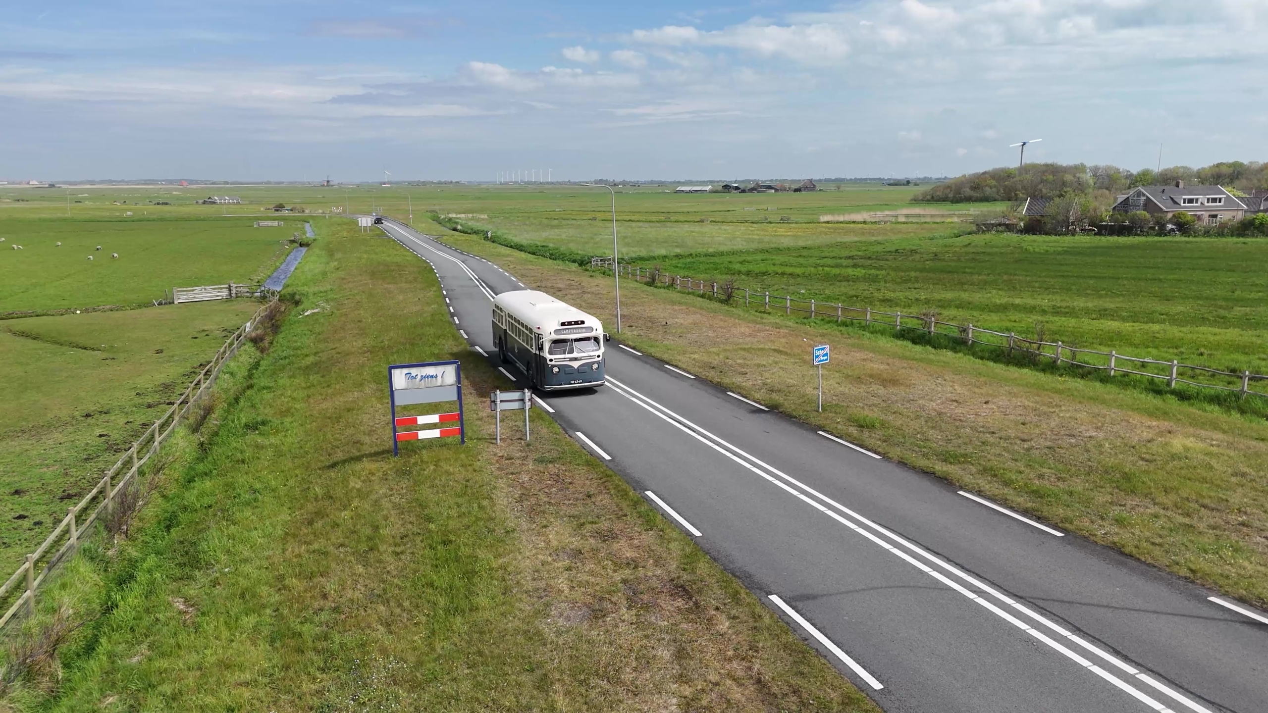 Een oude bus rijdt over een rustige weg door een landelijk gebied met groene velden en een paar windmolens in de verte.