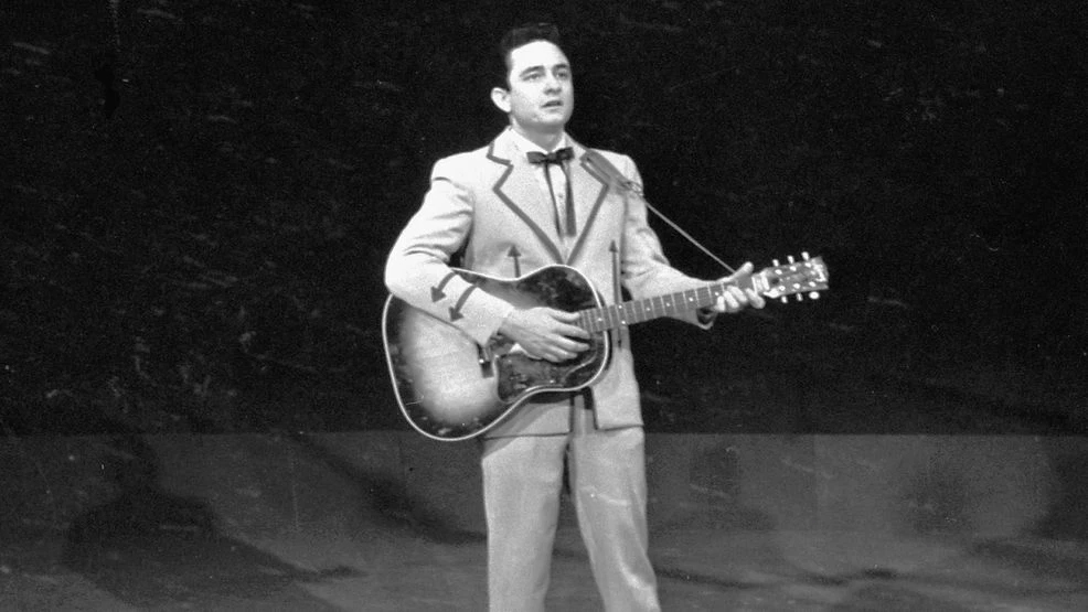 Een man in een pak speelt gitaar op een podium in een zwart-witfoto.