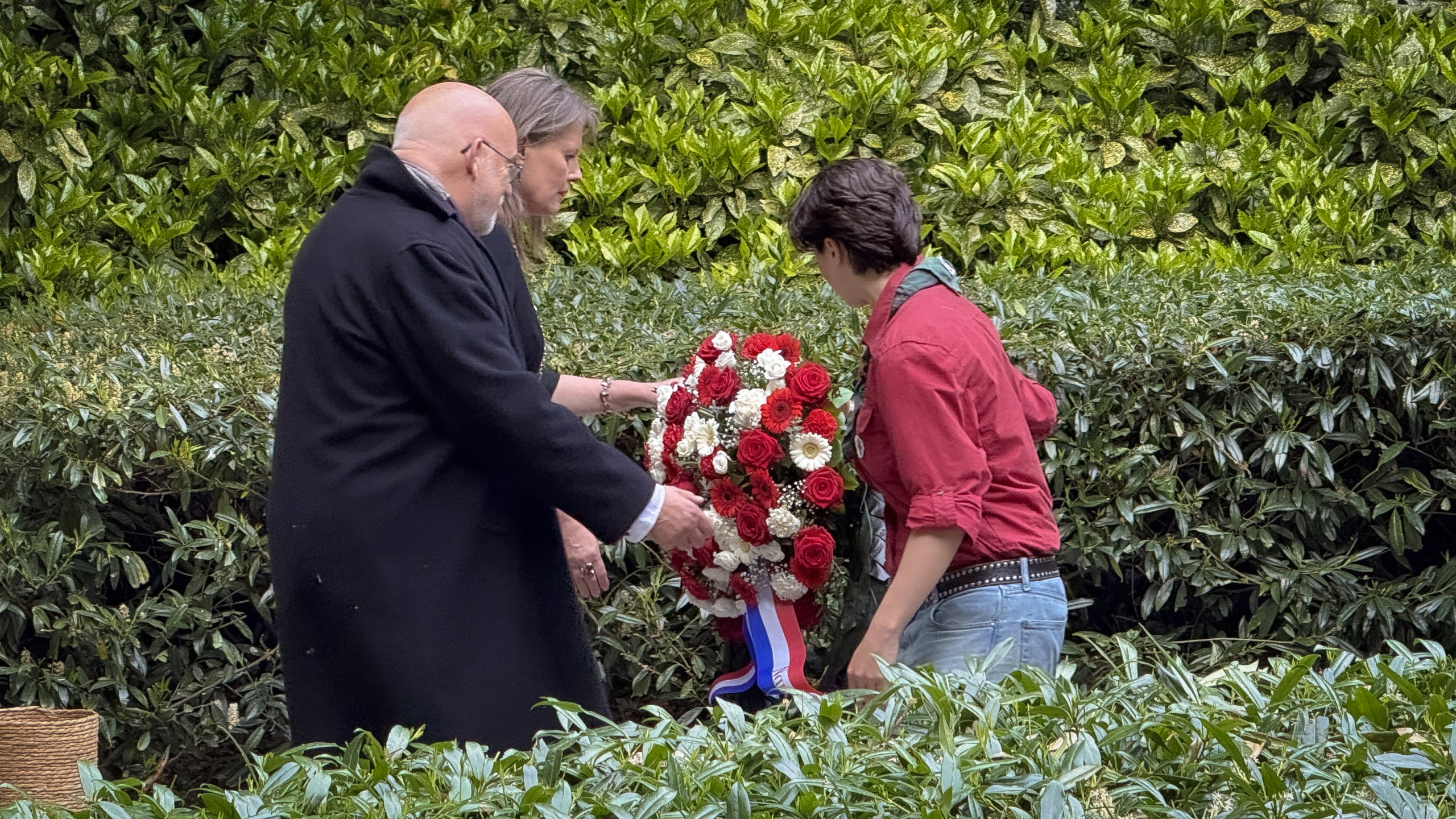 Drie mensen leggen een krans van rode en witte bloemen met een strik in rood-wit-blauw voor een groene heg.