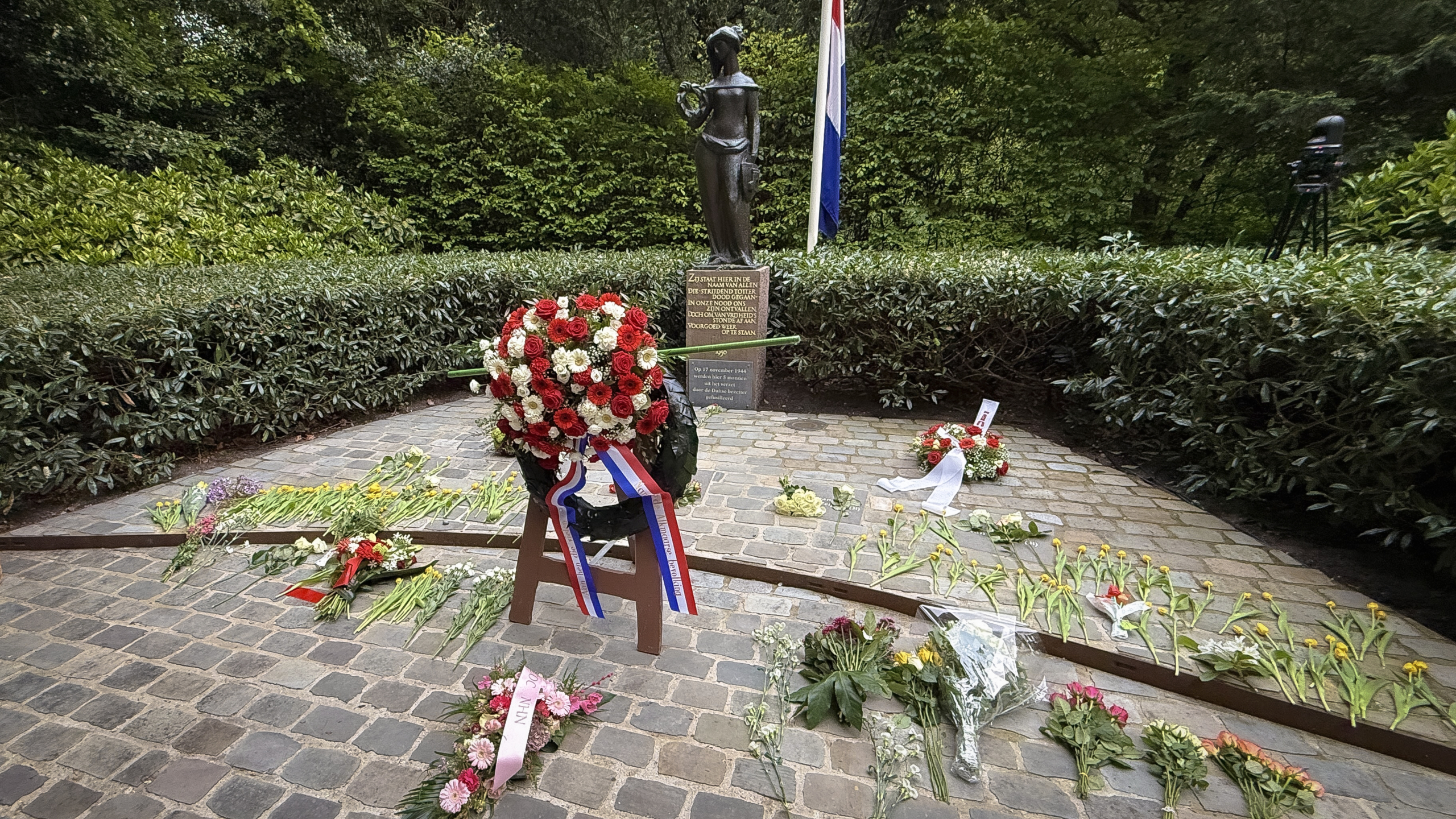 Bloemenkransen en -boeketten liggen bij een standbeeld en een plaquette, omgeven door groen, met daarnaast een Nederlandse vlag.