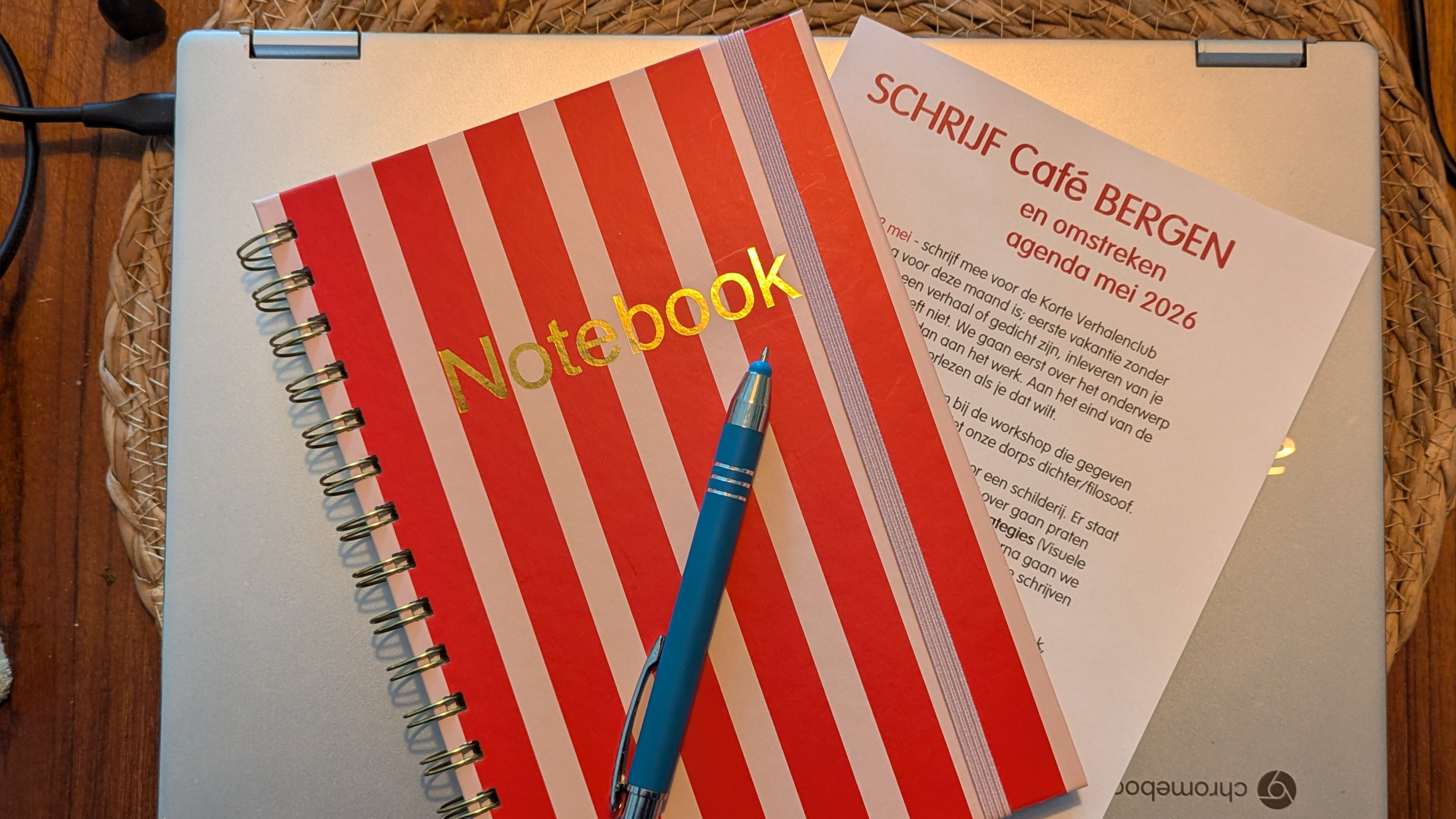 Rood-wit gestreept notitieboek en blauwe pen liggen op een laptop met een flyer voor 'SCHRIJF Café BERGEN' agenda mei 2026.