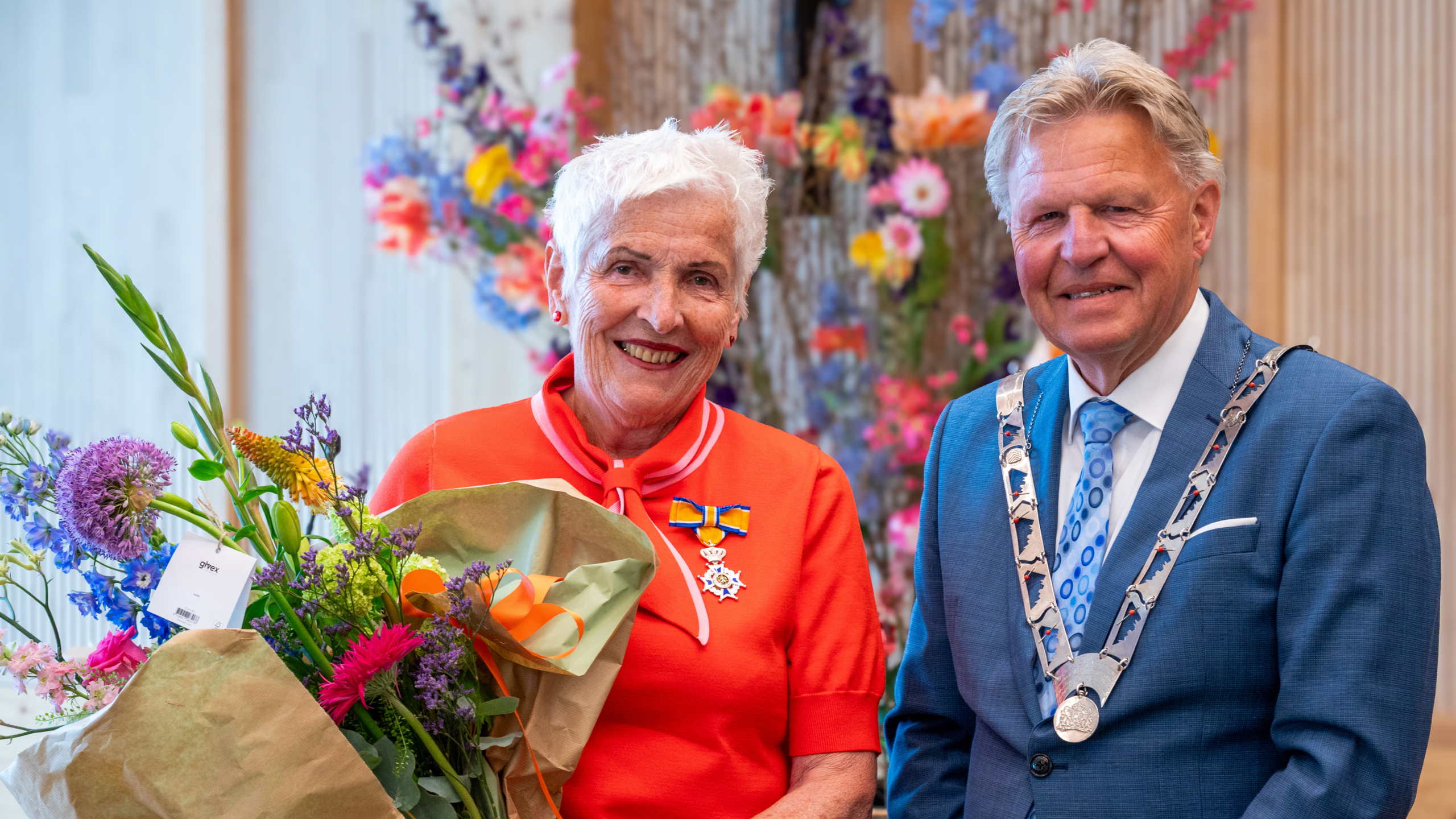 Een glimlachende vrouw met een koninklijke onderscheiding ontvangt bloemen naast een man in een blauw pak met ambtsketen.