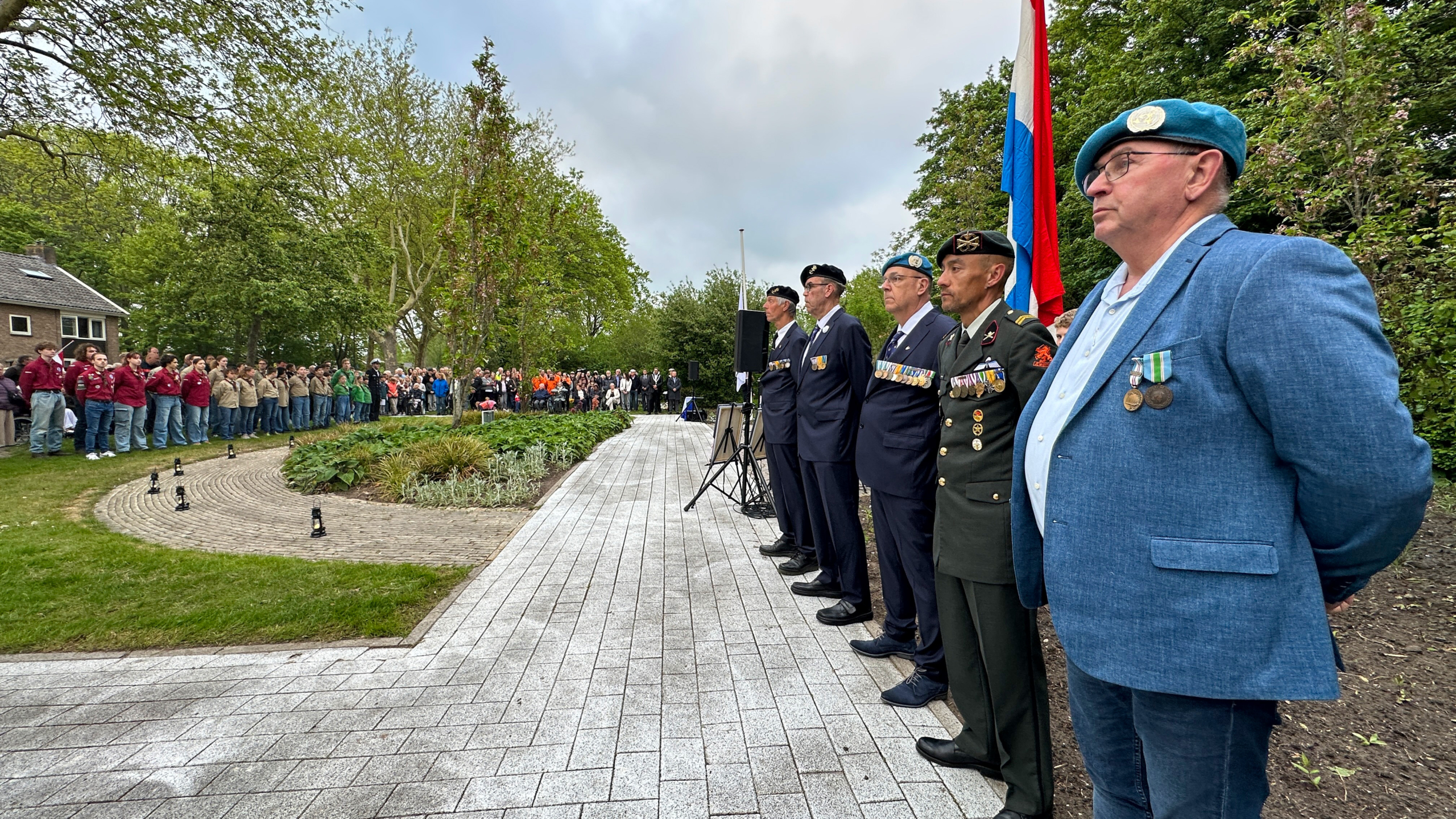 Veteranen en scouts staan in formatie bij een herdenkingsceremonie, met een Nederlandse vlag op de achtergrond.