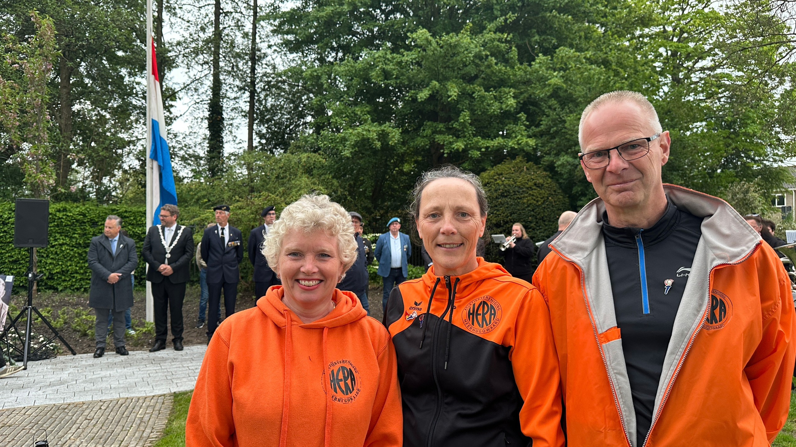 Drie mensen in oranje outfits staan voor een groep mensen bij een herdenking met een Nederlandse vlag halfstok.