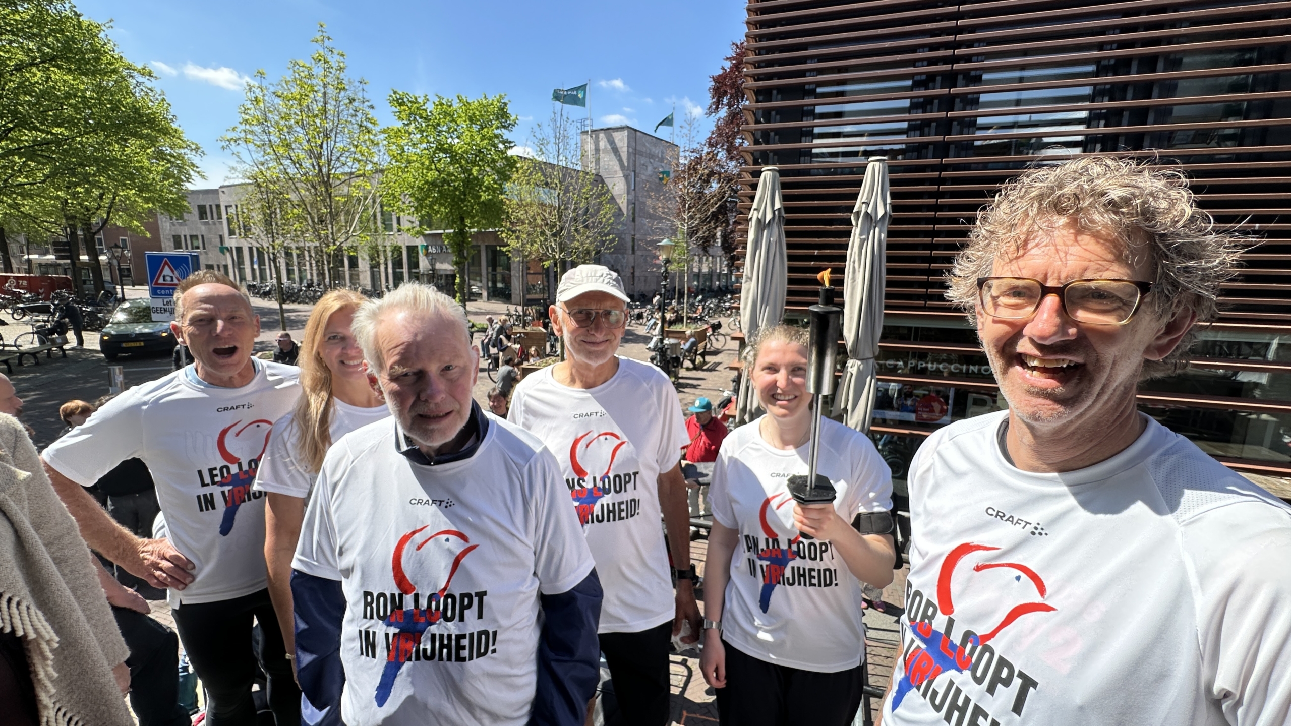 Een groep mensen in witte T-shirts met teksten verzameld buiten in de zon, met een vrouw die een fakkel vasthoudt.