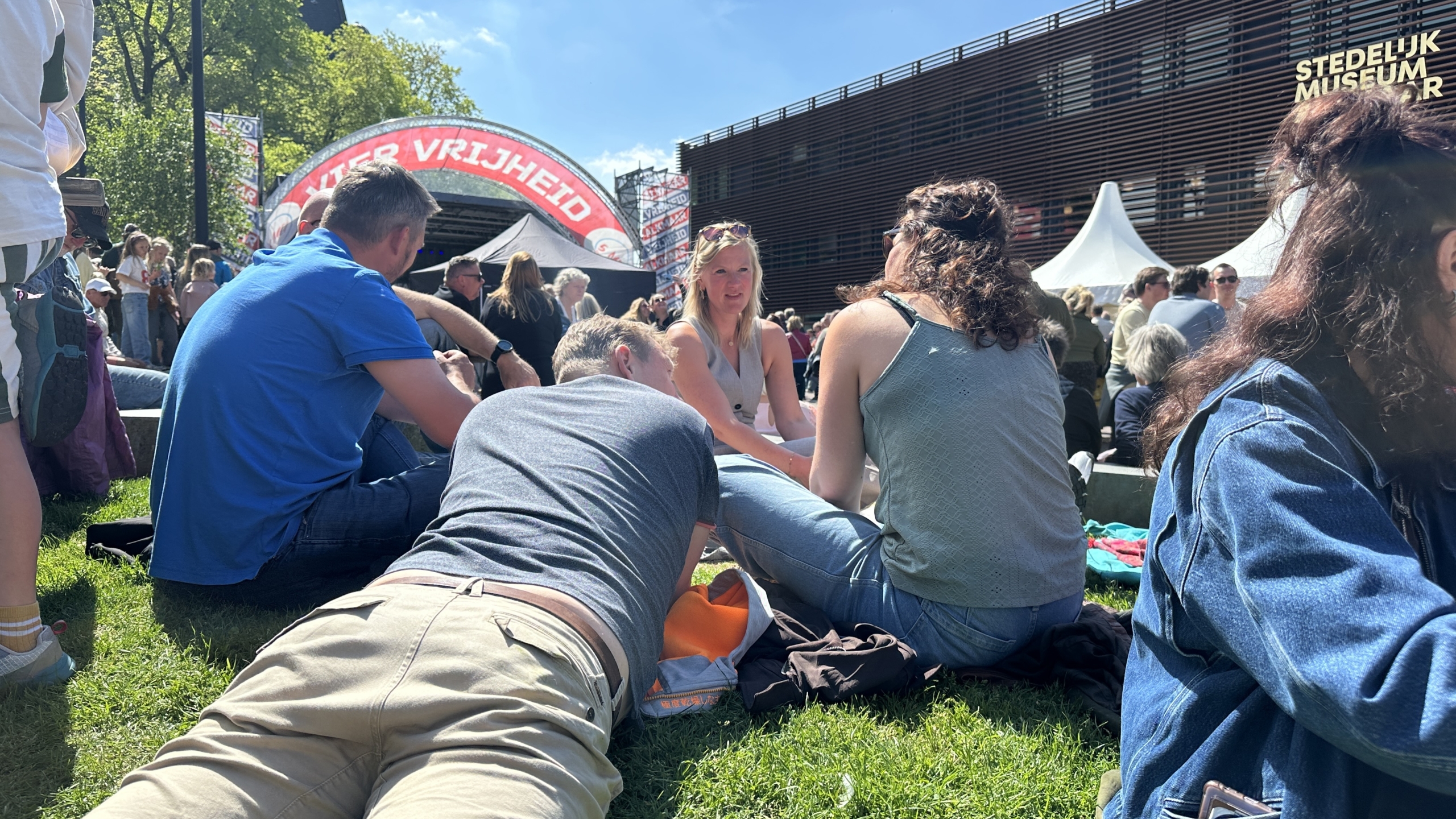 Groep mensen zit op het gras bij een evenement, met een boog met de tekst "Vier Vrijheid" en het Stedelijk Museum op de achtergrond.