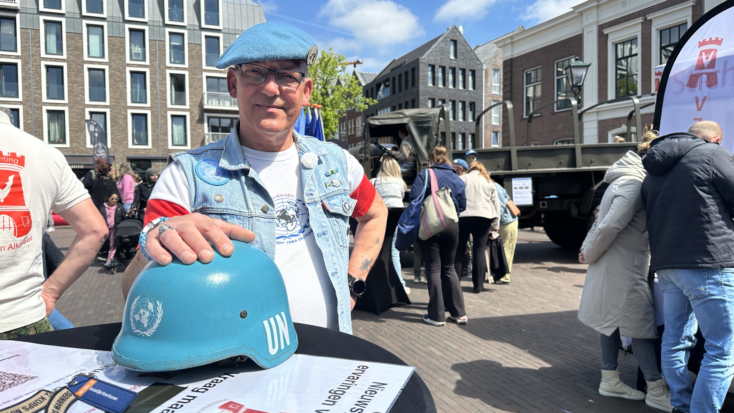Man met blauwe UN-helm en baret staat buiten met een informatiestand, omringd door mensen en gebouwen.