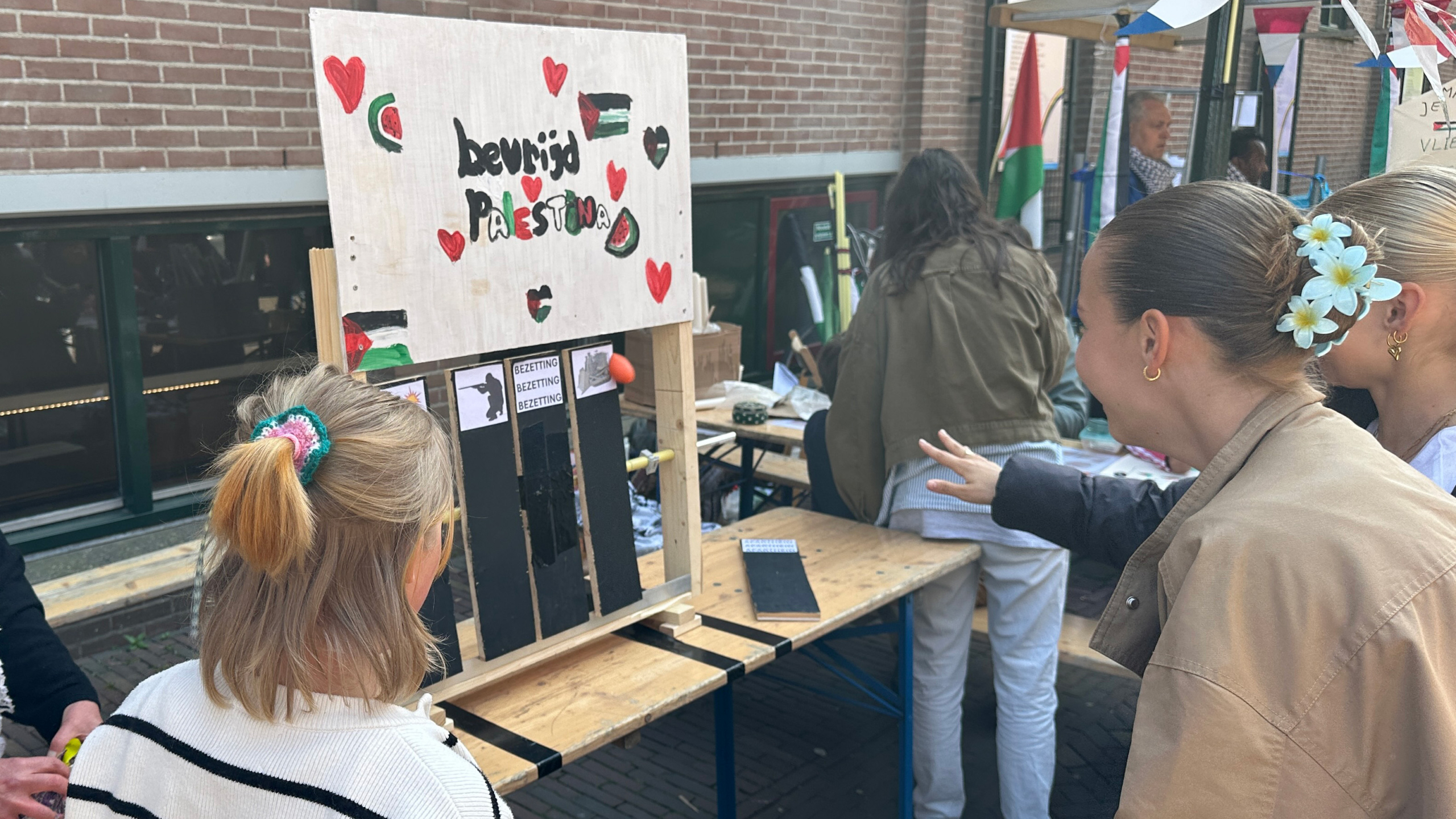 Kinderen kijken naar een houten bord met de tekst "bevrijd Palestina" en versieringen, tijdens een marktevenement.