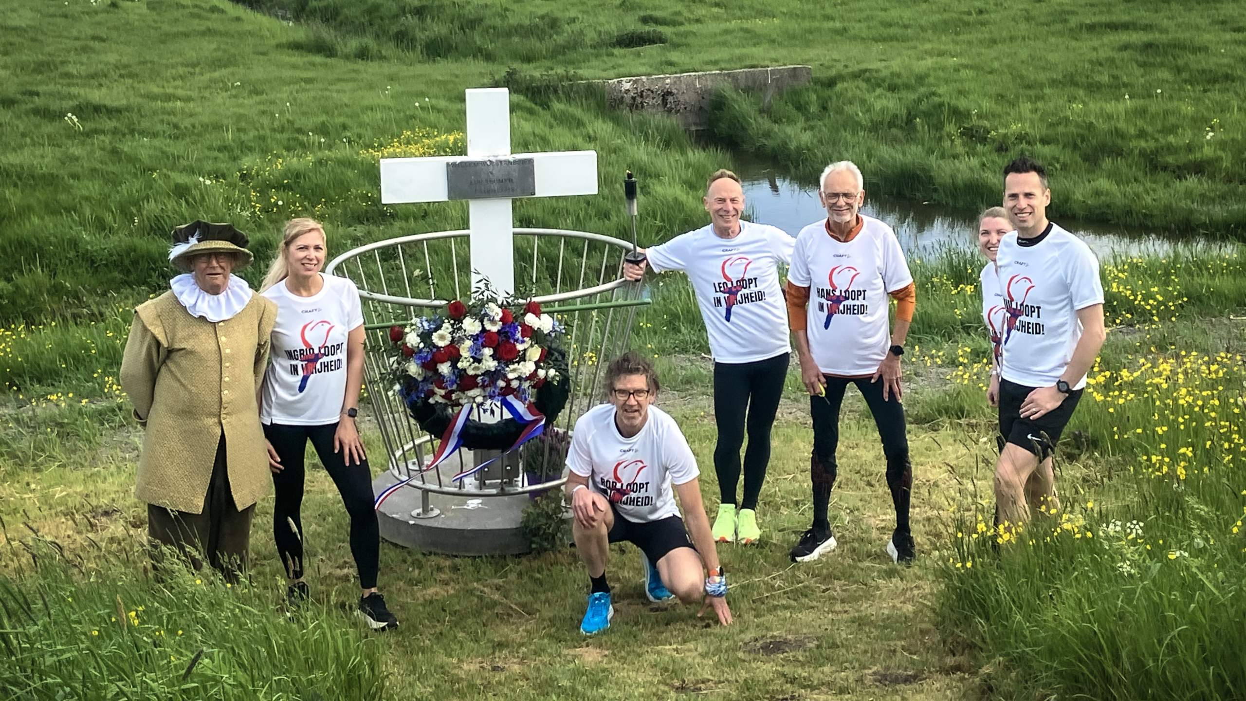 Een groep mensen in sportkleding staat bij een herdenkingsmonument met een groot wit kruis en bloemen, op een grasveld.