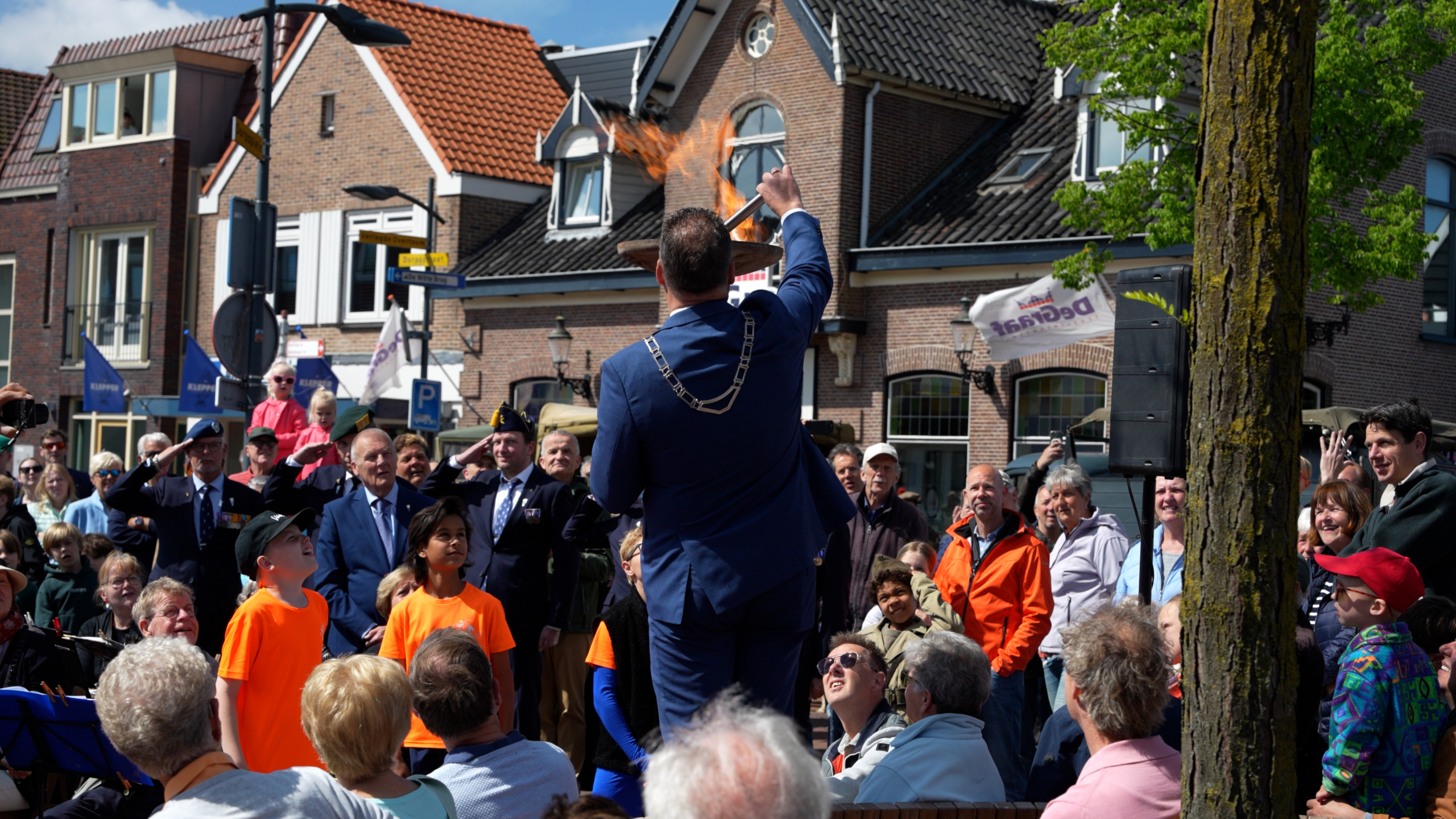 Een man in een blauw pak met een ambtsketen houdt een brandend vuur vast voor een publiek in een levendige straat.