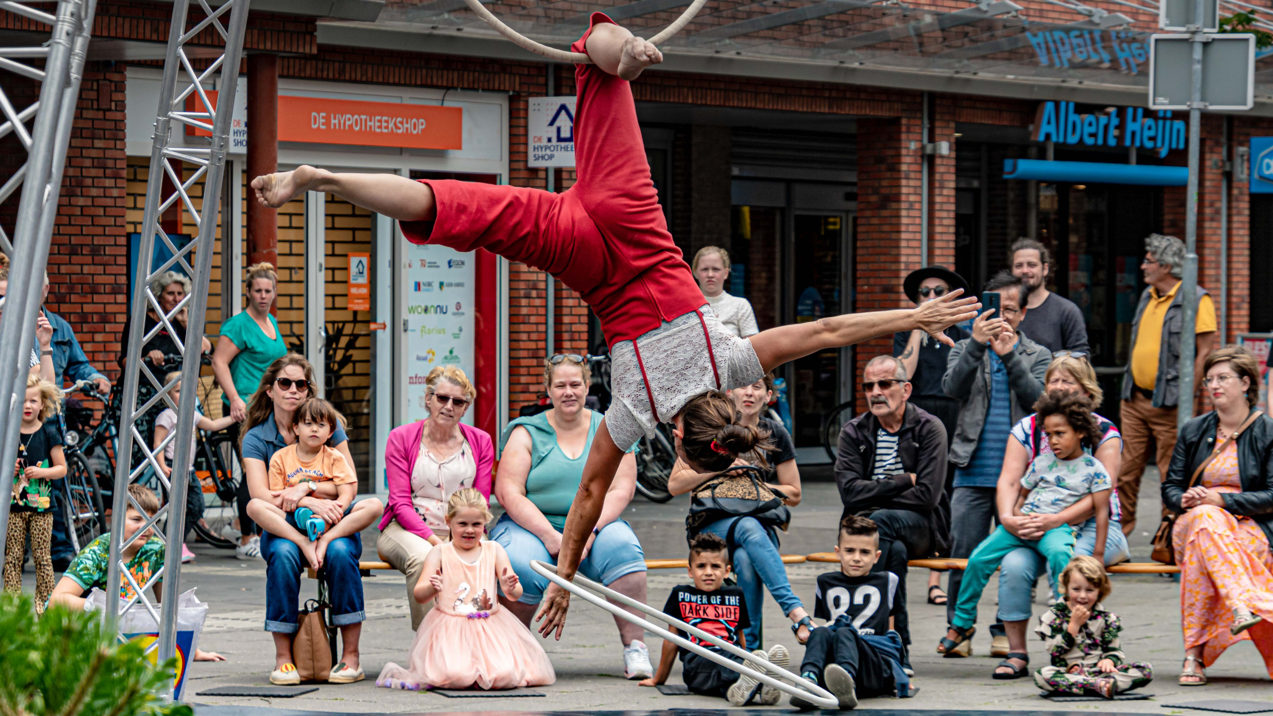 Een acrobaat in een rode outfit voert een truc uit aan een ring, voor een publiek op straat.