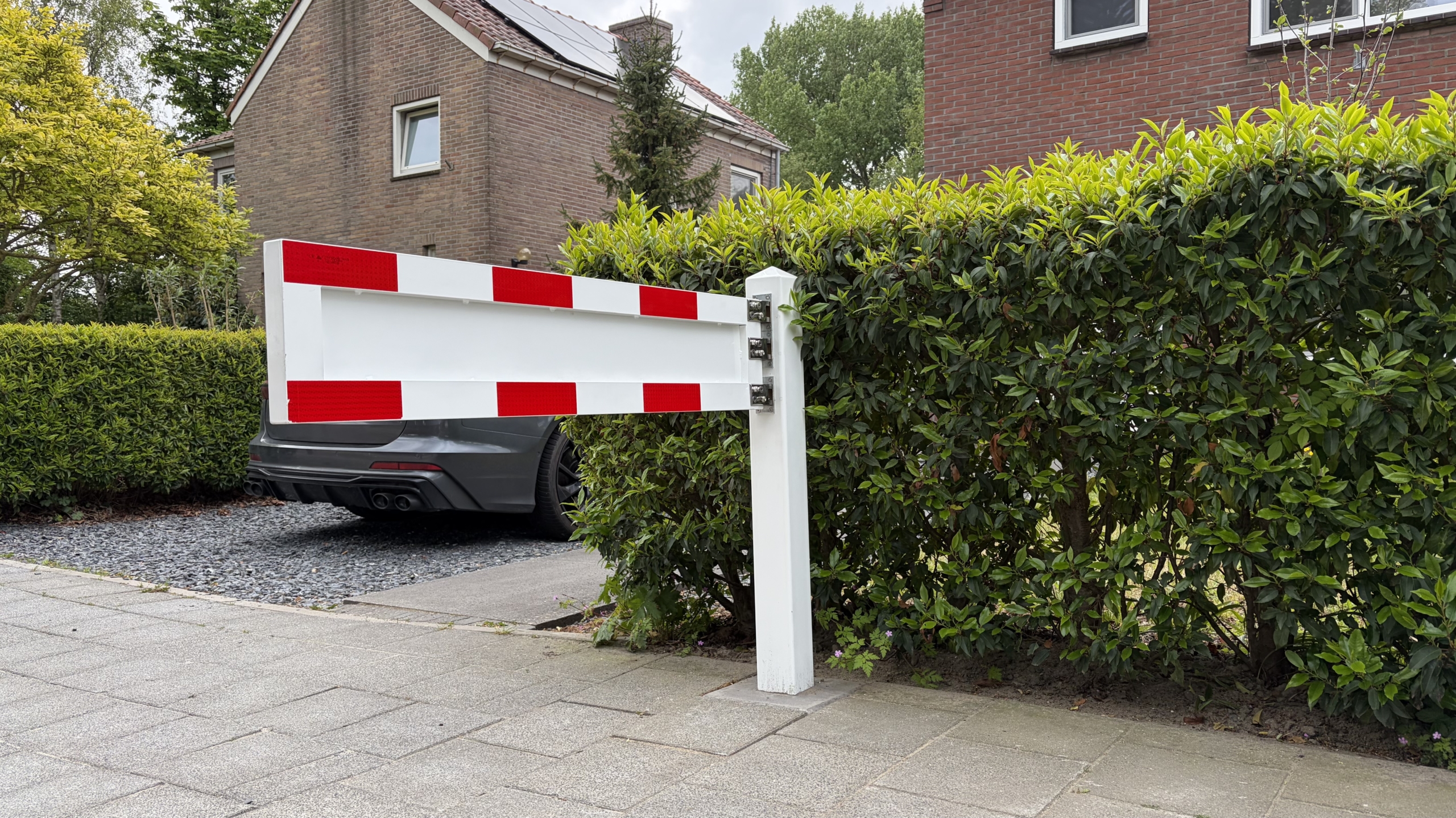 Wit-rood slagboom naast heg bij oprit van een bakstenen huis, met een geparkeerde auto erachter.