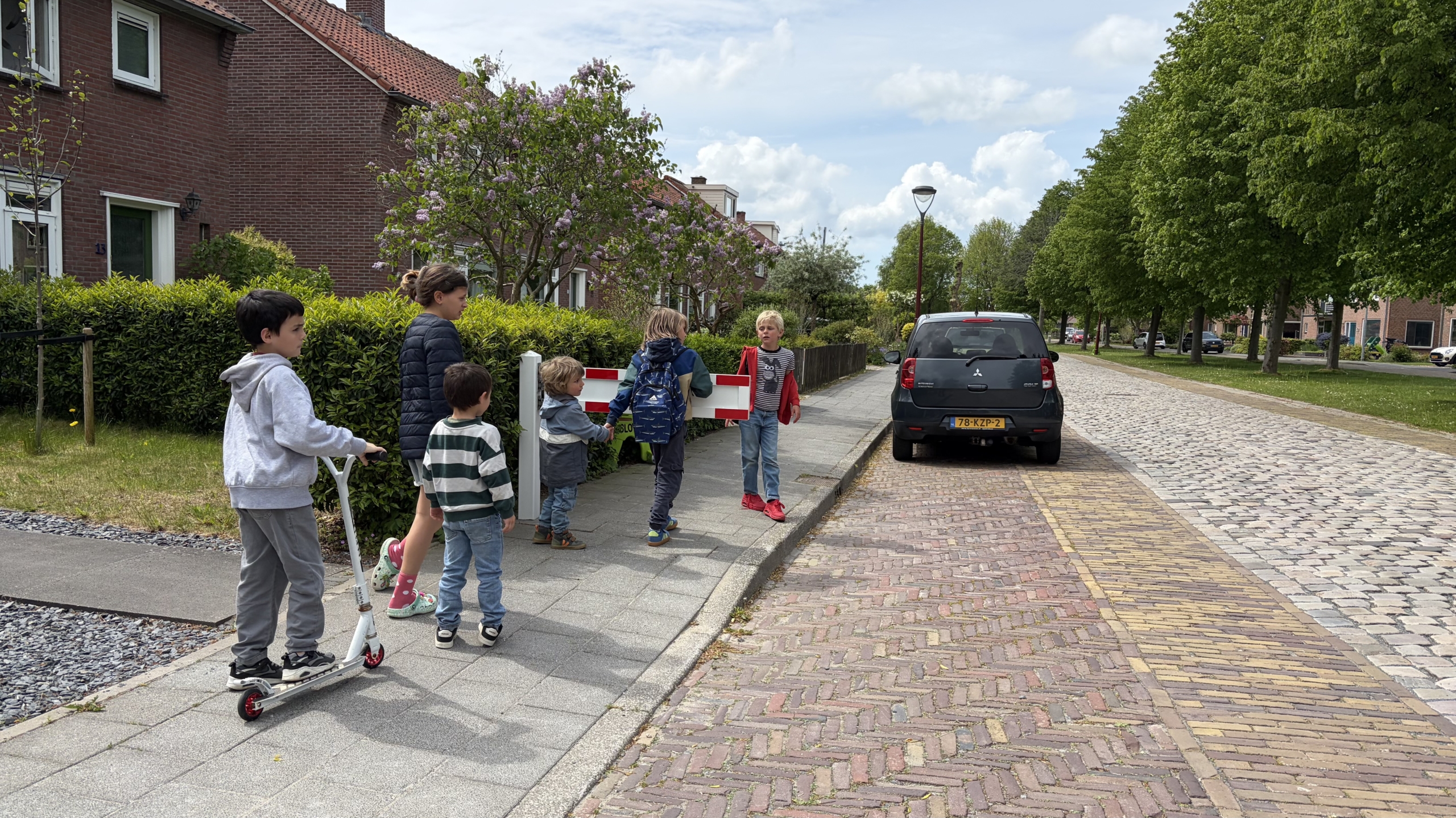 Kinderen en een vrouw staan op een stoep naast een auto en een hek, omringd door huizen en groene bomen.