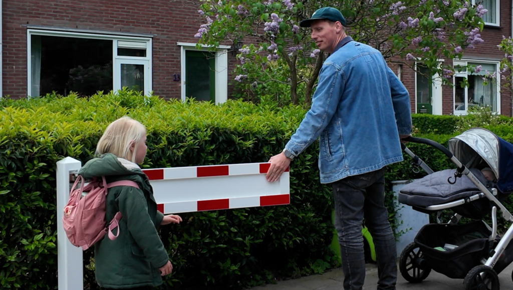 Man en kind bij een rood-witte afsluiting voor een huis, met een kinderwagen en bloeiende struik op de achtergrond.