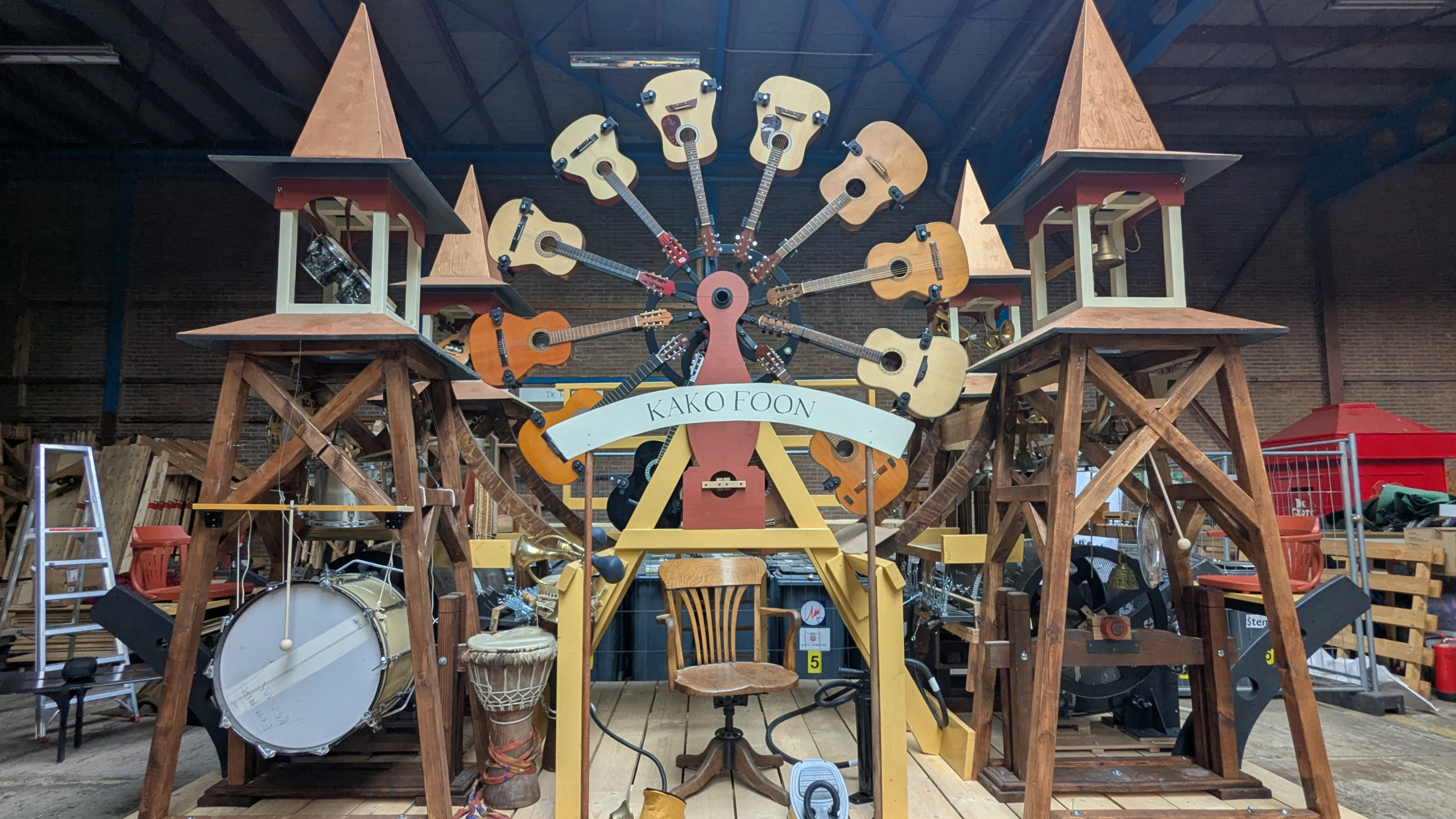 Een muziekinstallatie met gitaren geplaatst op een wiel, geflankeerd door houten torens, met de tekst "Kako Foon".
