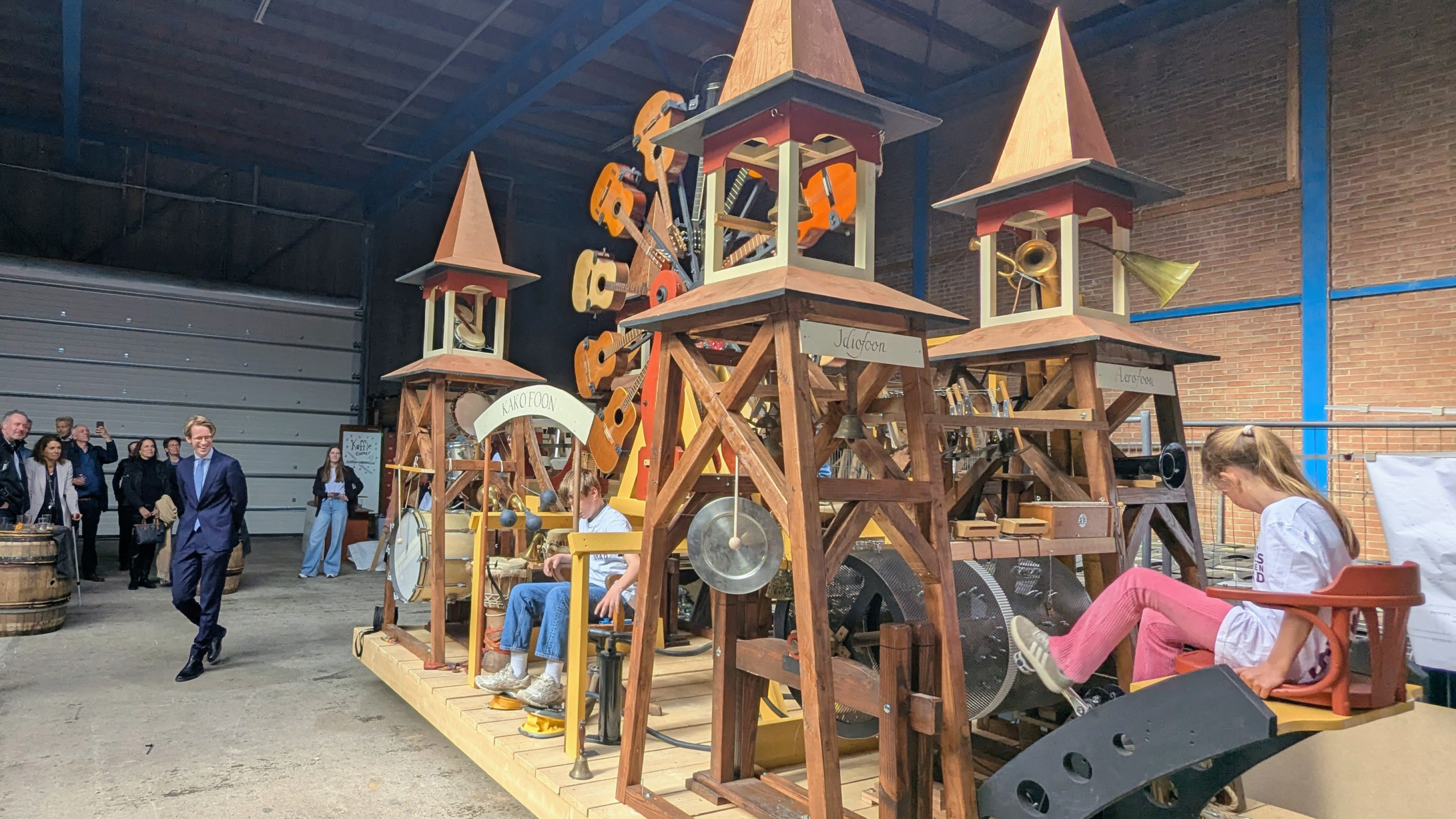 Een grote, houten machine met muziekinstrumenten en torenspitsen, bediend door kinderen; toeschouwers kijken toe.