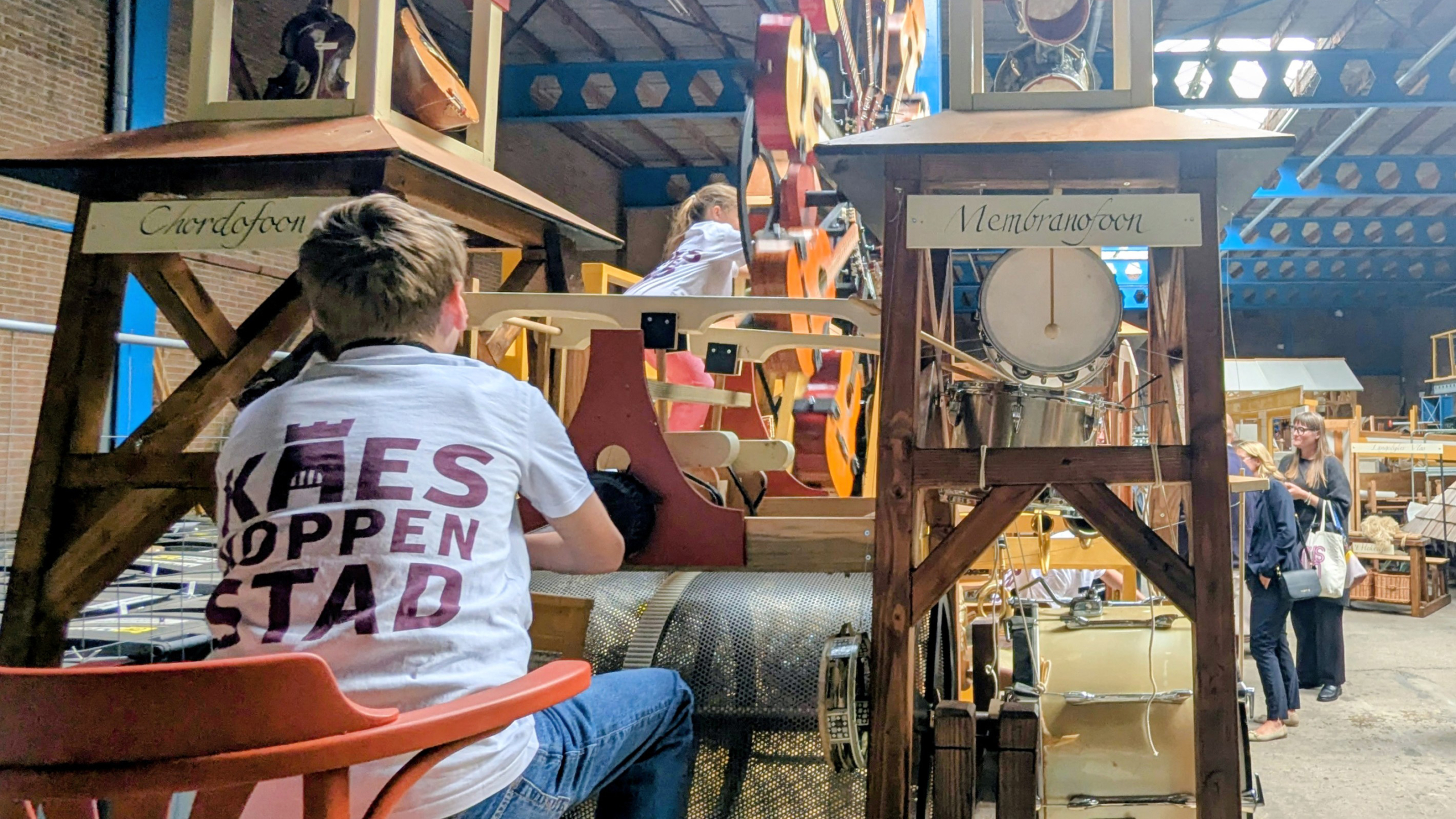 Twee kinderen zijn bezig met een groot houten muziekmechanisme in een werkplaats, omringd door verschillende muziekinstrumenten.