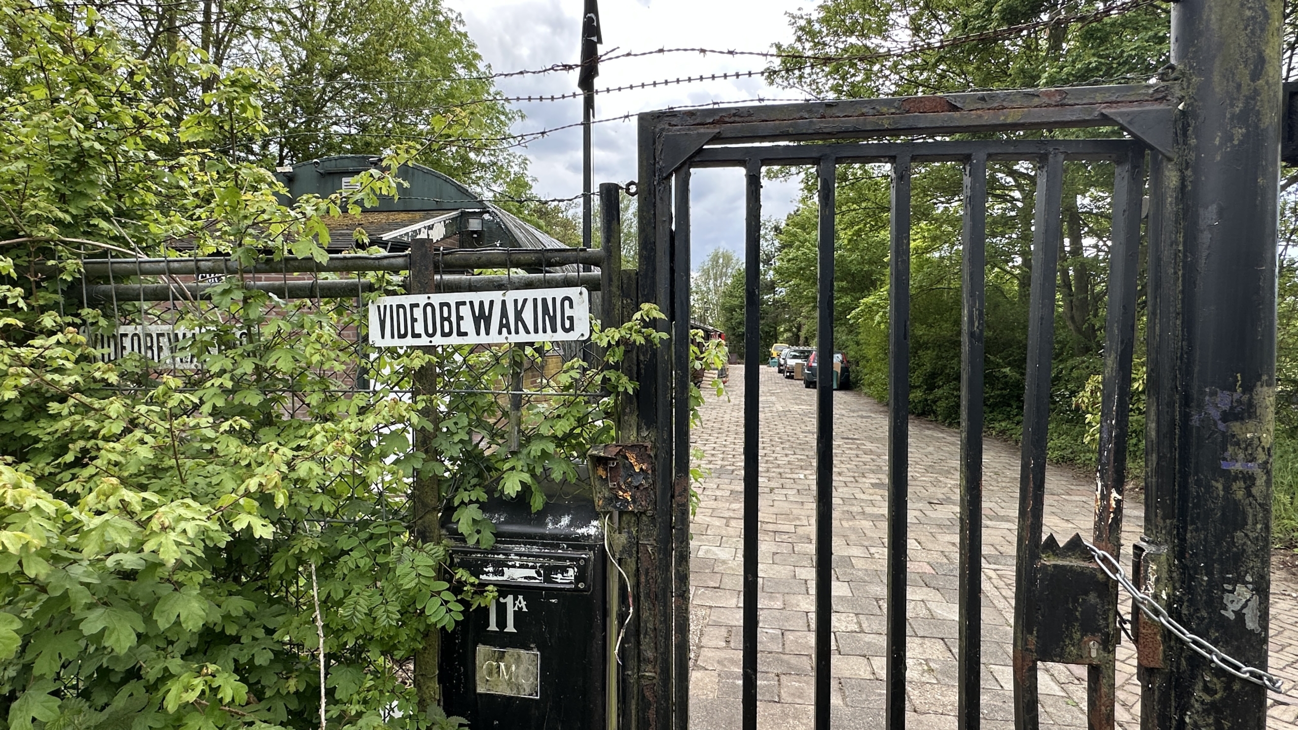 Openstaande ijzeren poort met bord "videobewaking", omgeven door bladeren, leidt naar een weg met geparkeerde auto's.